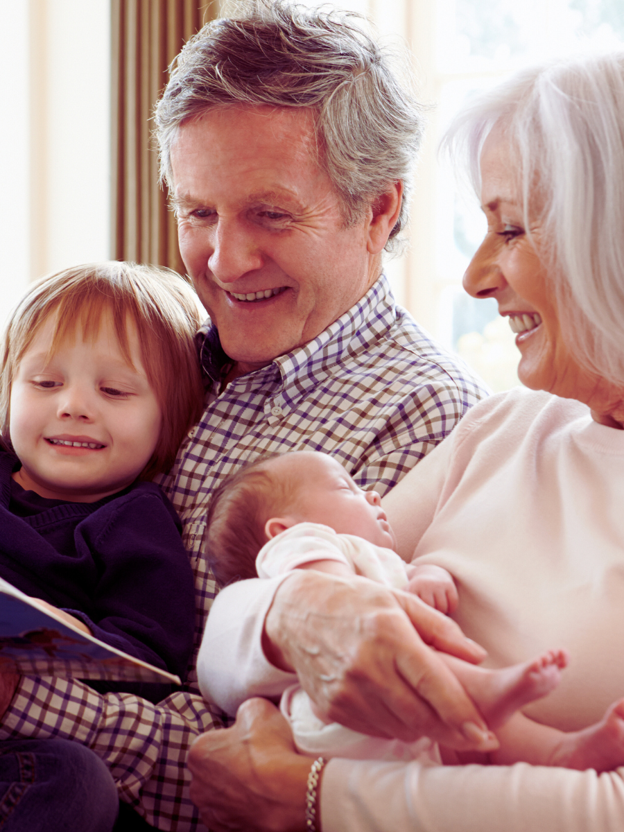 Grandparents with two grandchildren, one in the grandmother's arms and one sitting on the grandfather's lap, smiling and looking at a book. The grandparents are thinking about IHT and estate planning
