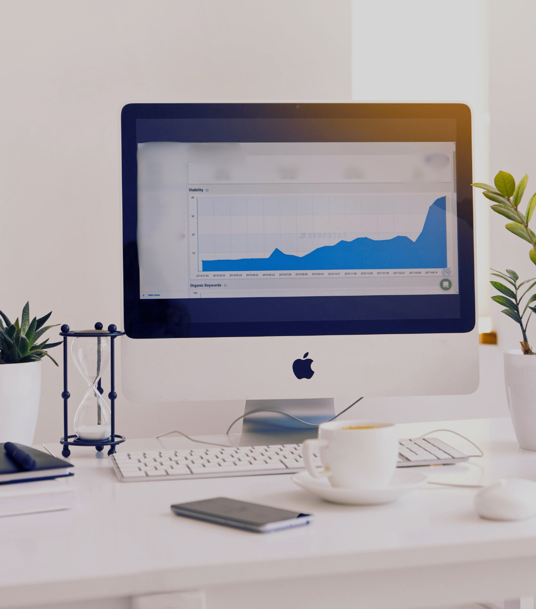 Apple iMac computer on a white desk displaying a line graph showing an upward trend in website visibility over time, with potted plants, a cup of coffee, a notebook, and an hourglass on the desk.