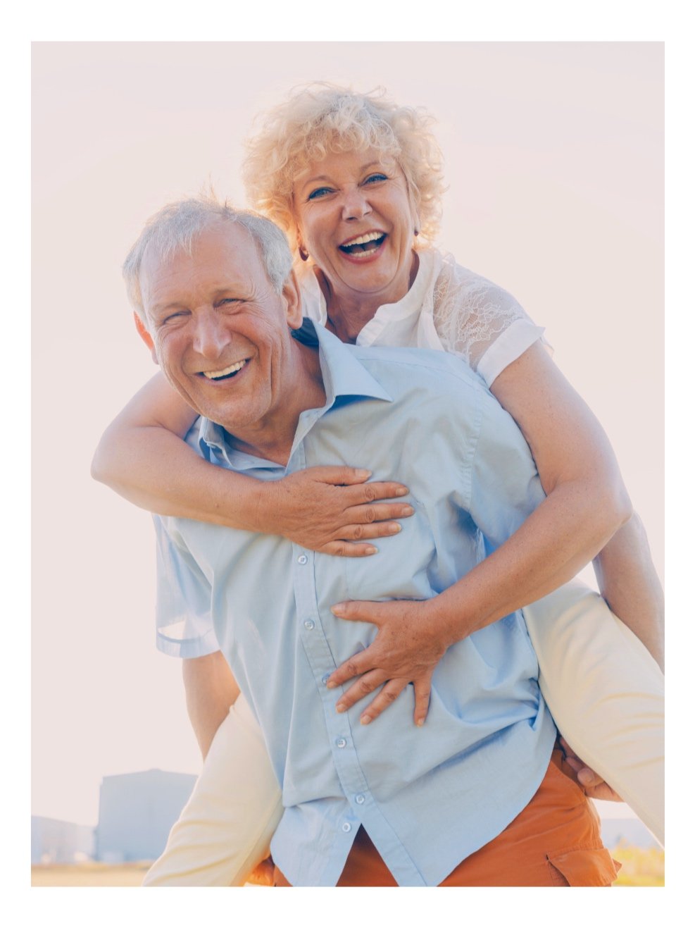 An elderly couple laughing and smiling happily, with the woman on the man's back during an outdoor scene.