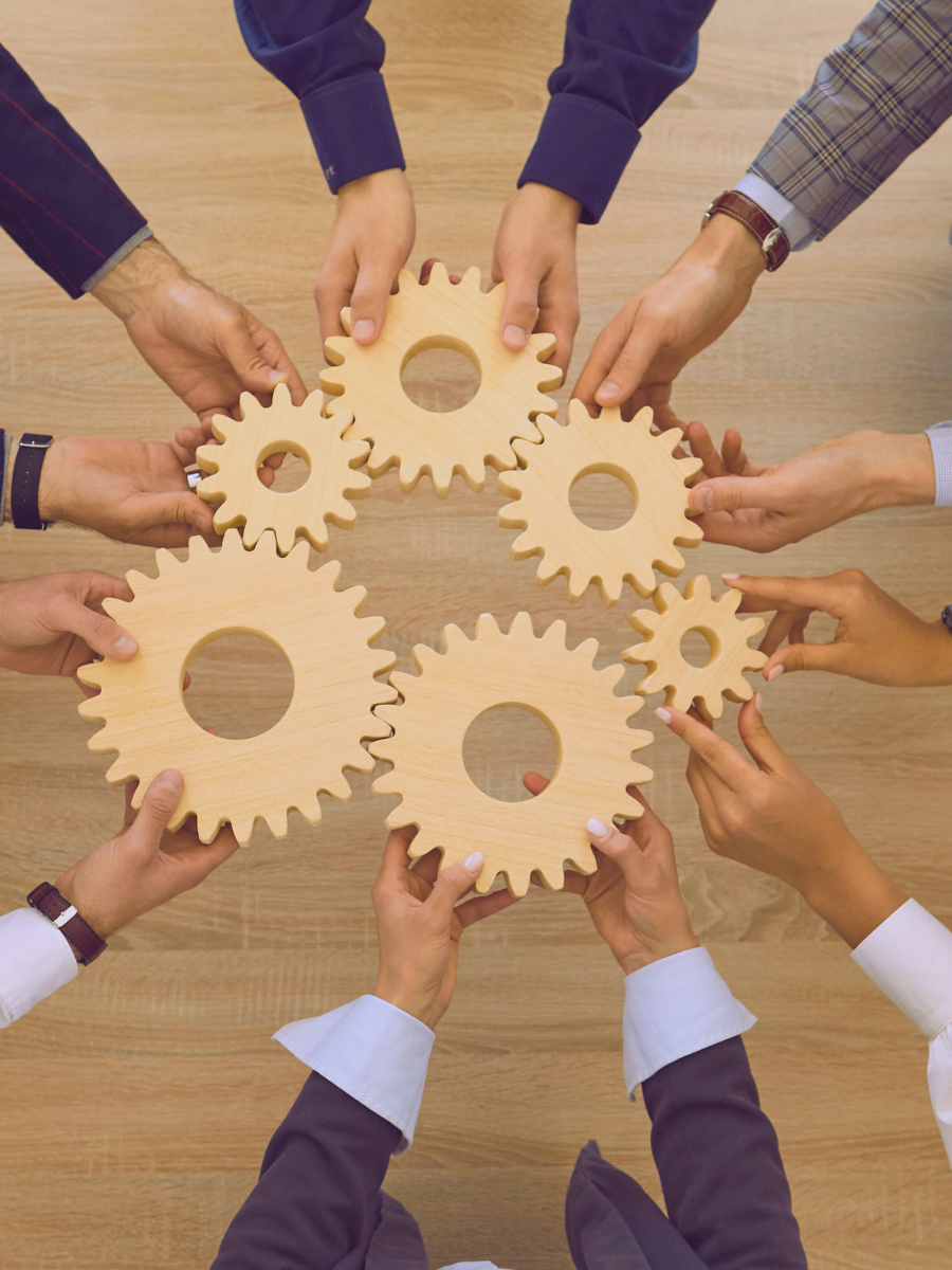 Six people holding interlocking wooden gears together on a wooden table.