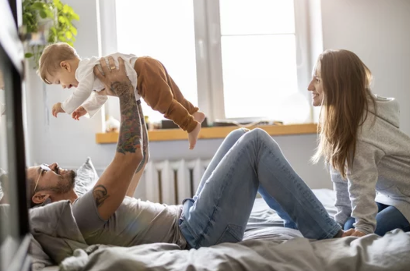 A family playing together on a bed in a sunny bedroom; a man lies on his back lifting a toddler into the air, a woman sits next to them smiling. They are considering life insurance needs and getting financial advice
