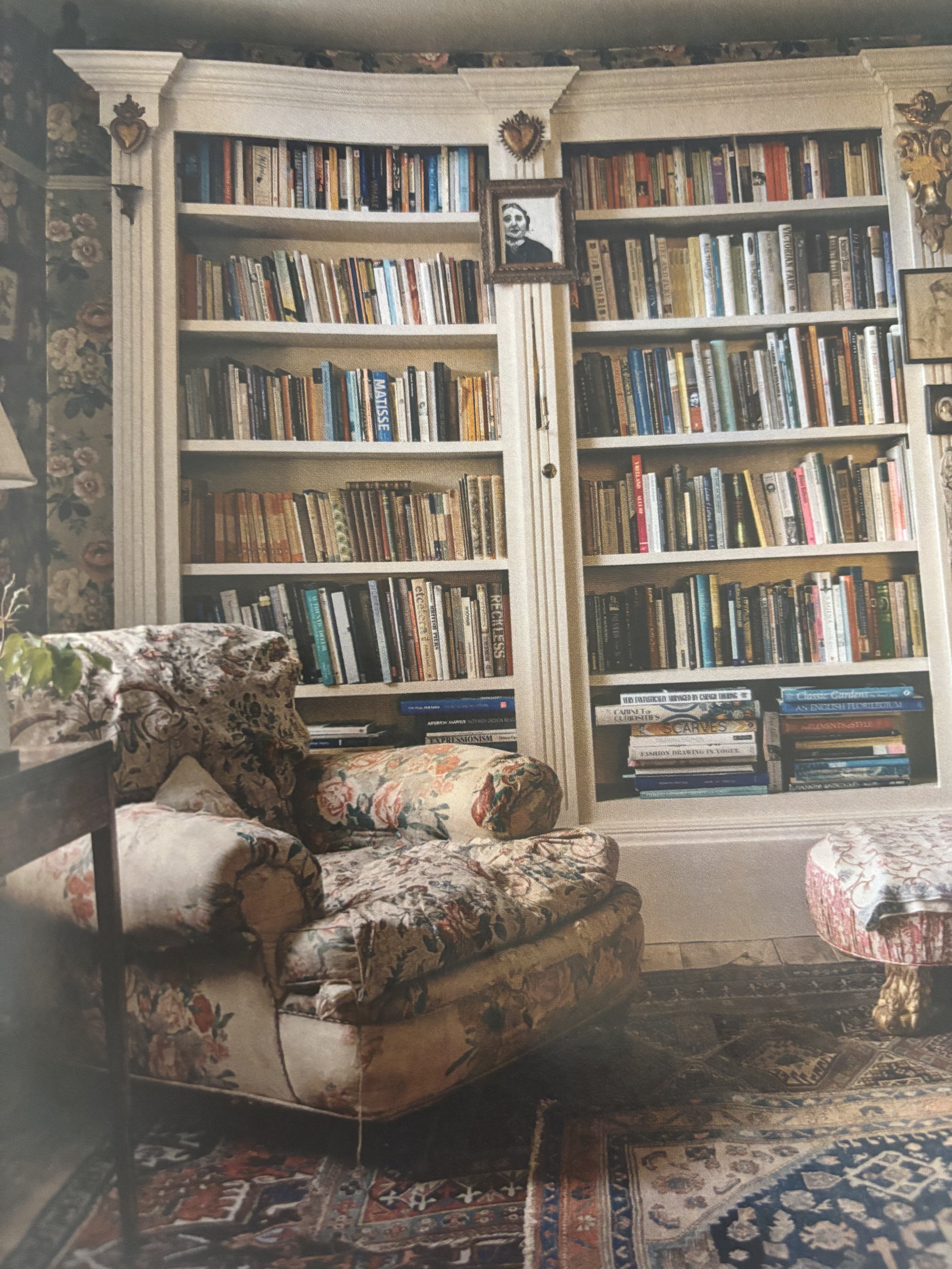 A cozy living room corner with a vintage floral patterned armchair, hardwood floor, and a large white bookshelf filled with books. There are framed pictures and decorative items on the shelves, and a Persian rug on the floor.