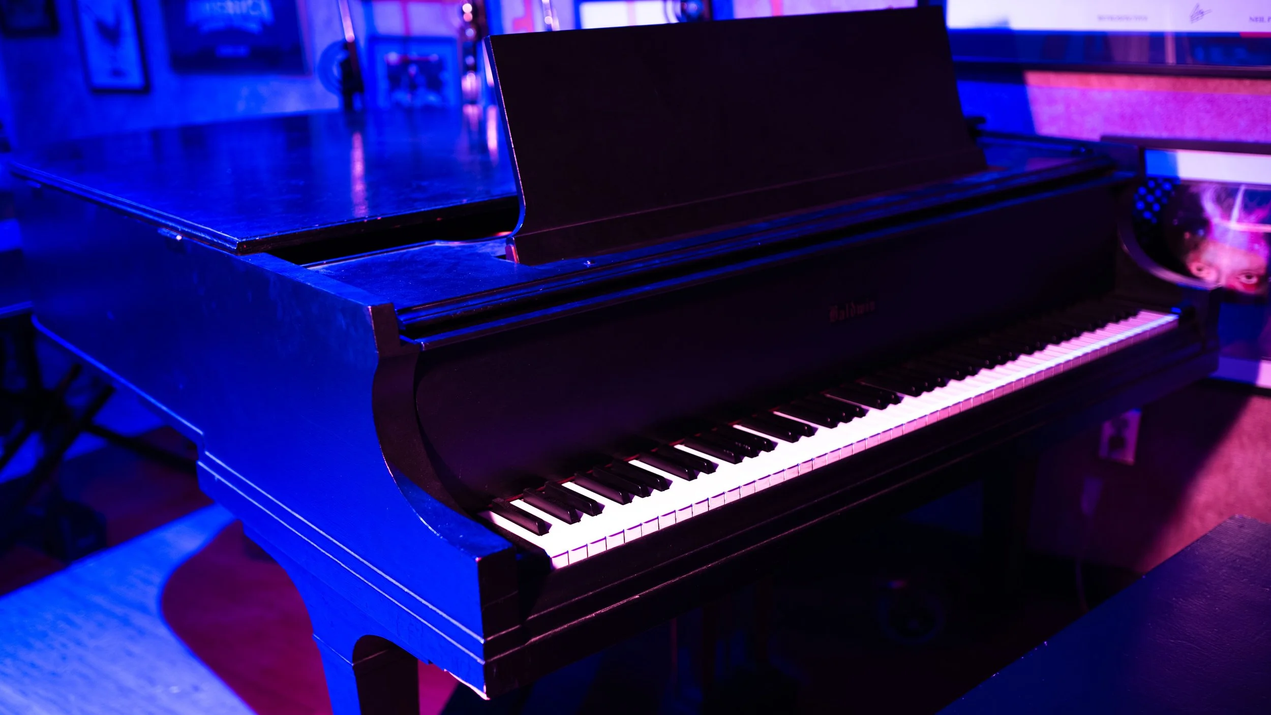 Black Baldwin piano in a dimly lit room with purple and pink neon lighting, a sheet music stand, and wall decorations.