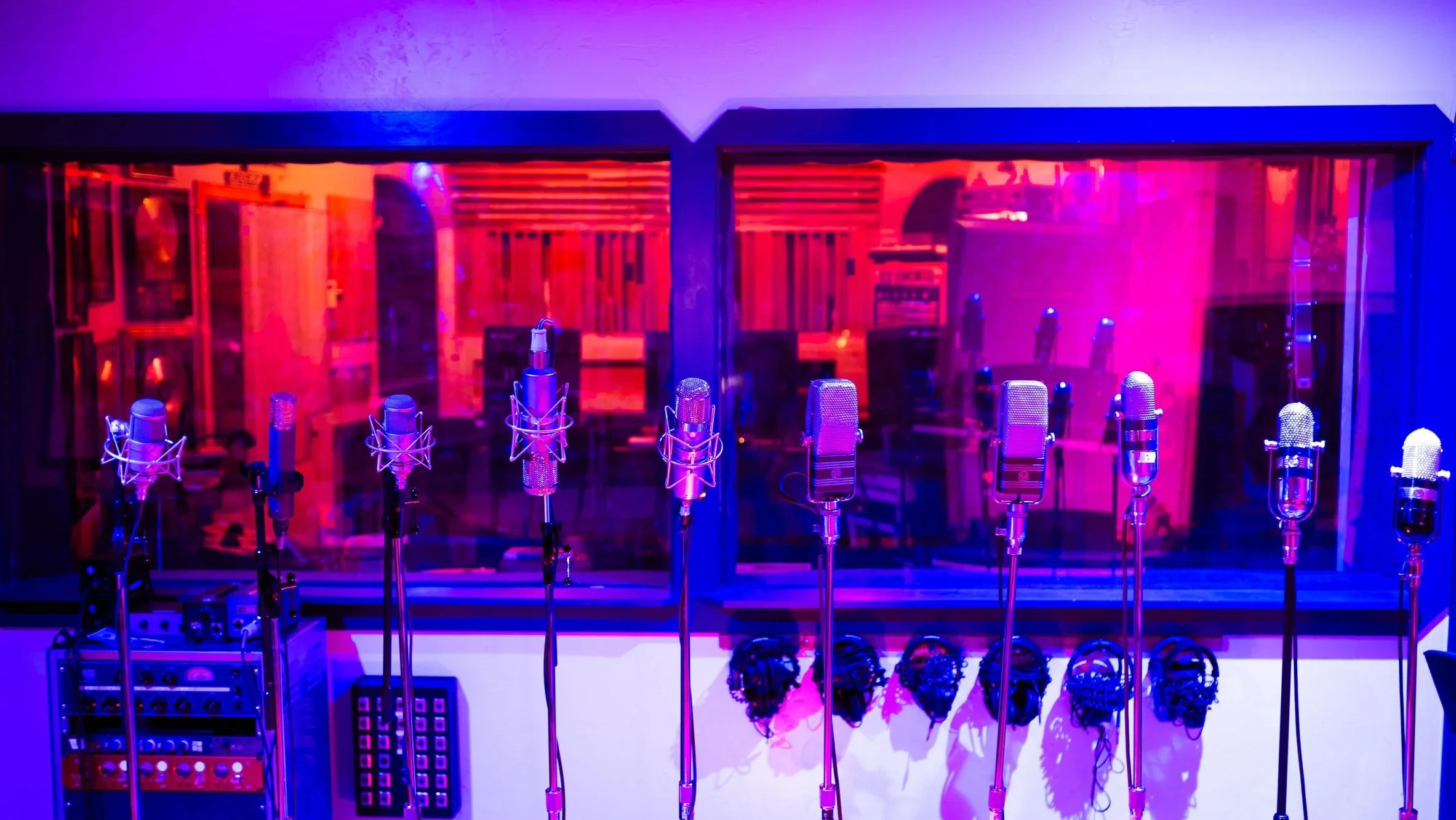Multiple microphones on stands arranged in a recording studio with colored purple and blue lighting and acoustic foam panels in the background.
