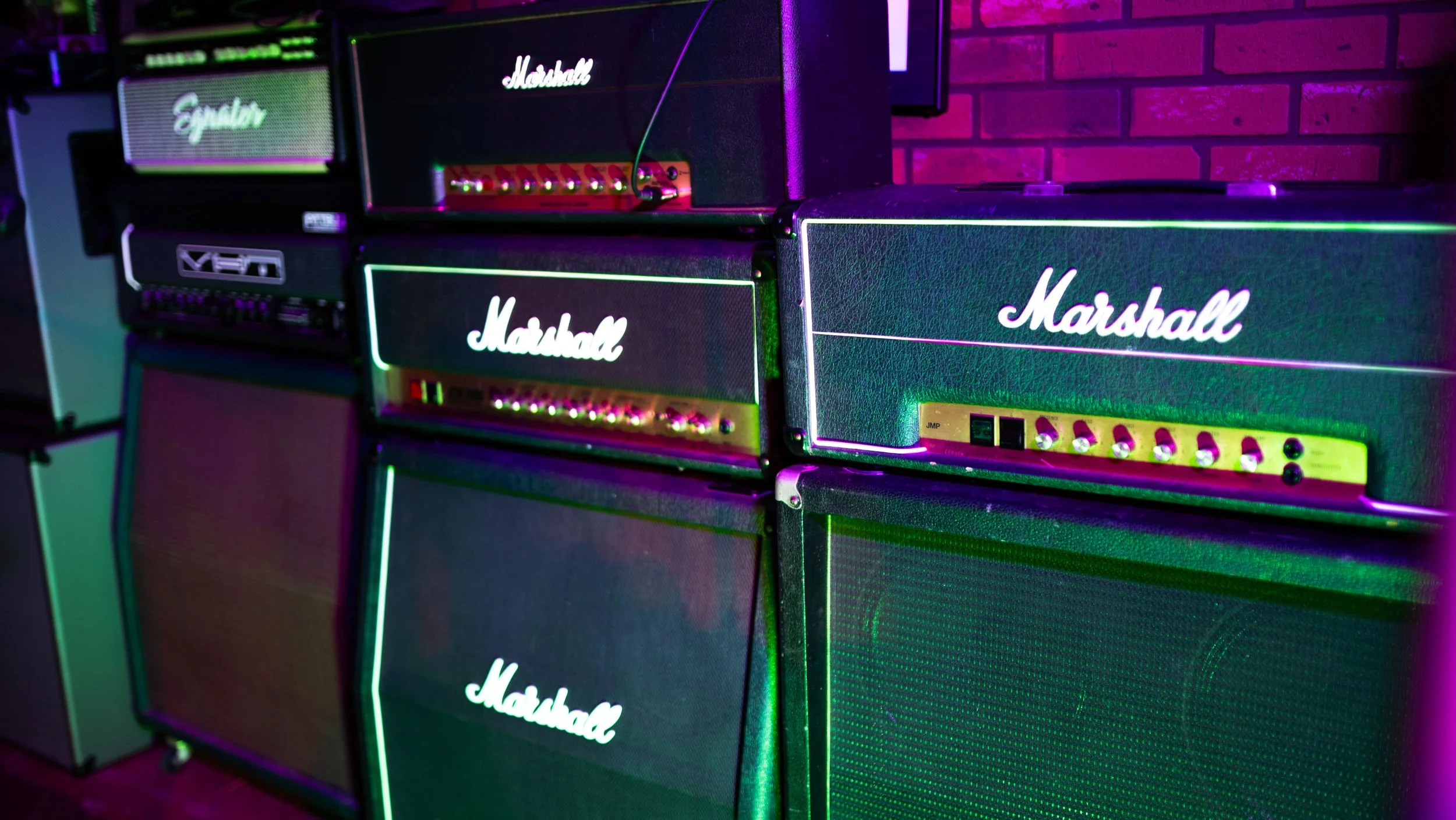 Stacked Marshall guitar amplifiers with colorful lighting and a brick wall background.