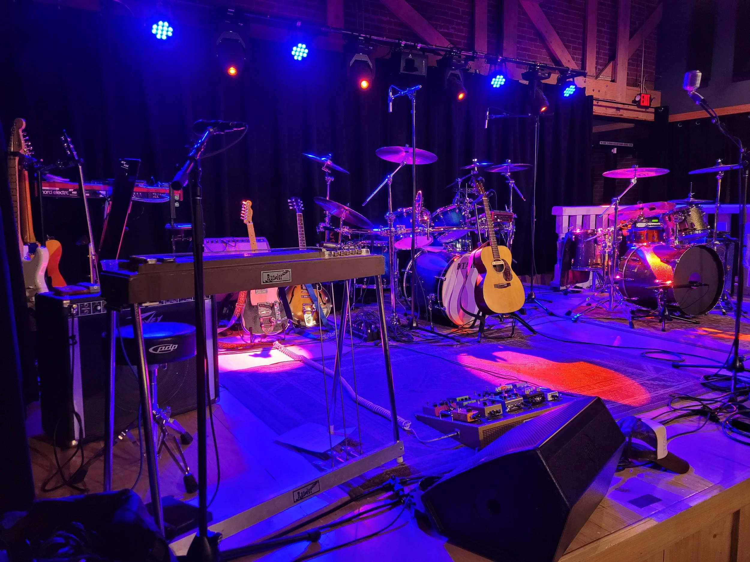 Empty musical stage with drums, guitars, keyboards, microphones, and various instruments under blue and purple stage lighting.