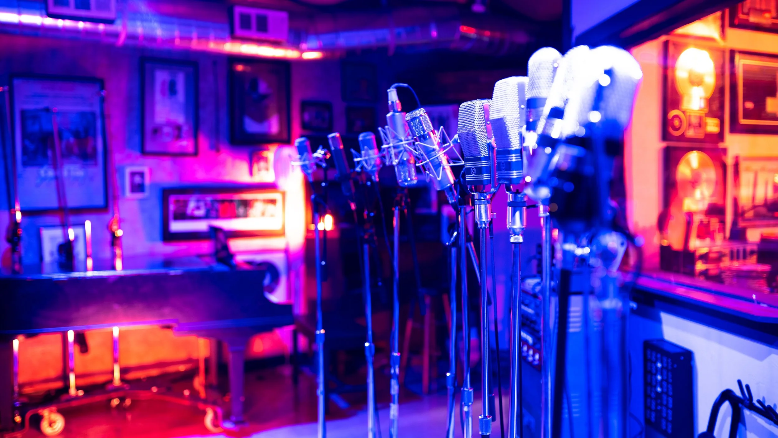 A row of microphones on stands in a music or recording studio with colorful pink and blue lighting, framed pictures on the wall, and musical equipment.