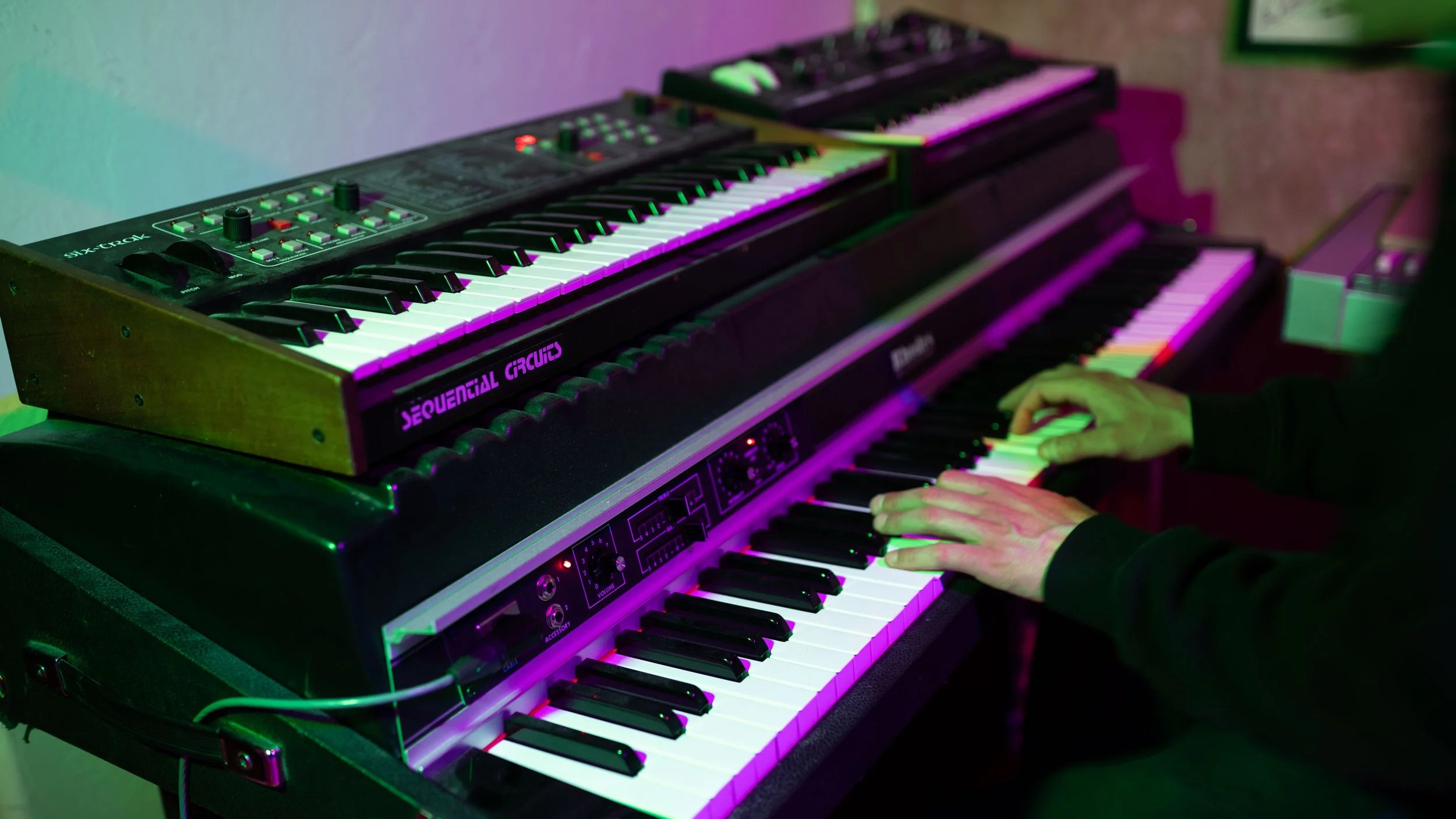 Person playing two layered synthesizers, one with black and white keys and the other with a digital interface, with colorful green and purple lighting effects.