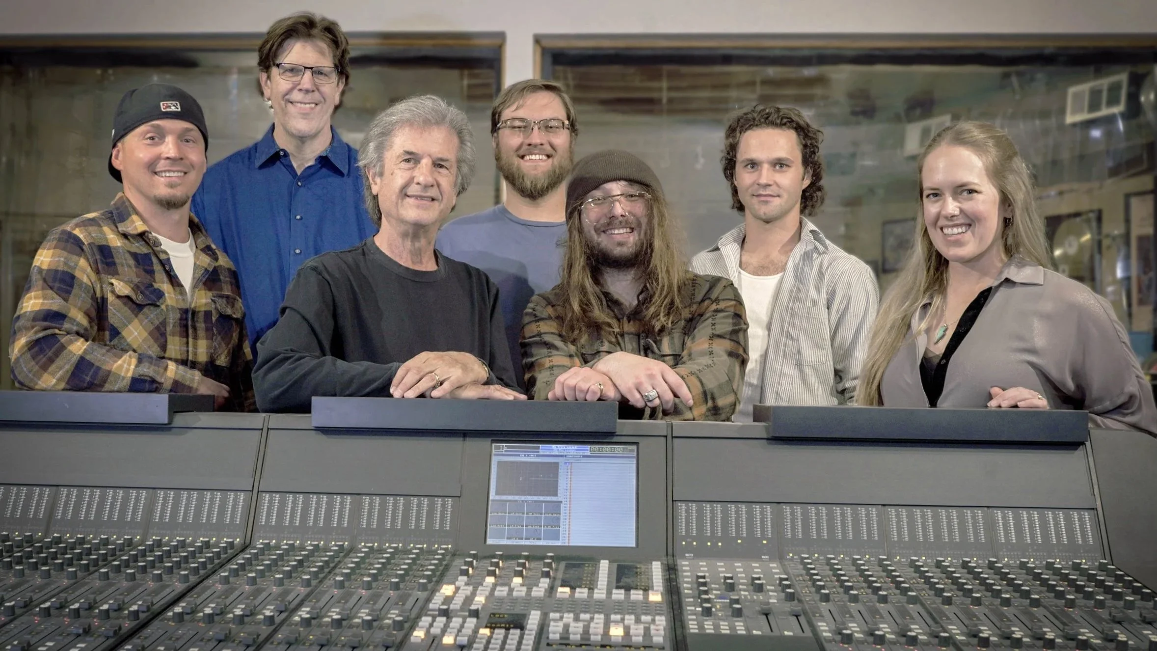Group of eight people standing behind an audio mixing console in a recording studio.
