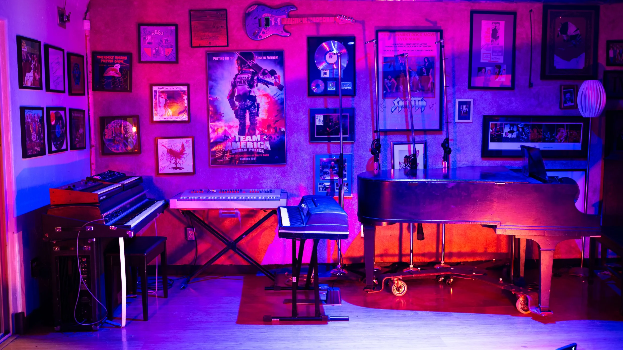 A music studio room decorated with posters on the walls, including a large 'Team America' poster. The room contains a grand piano, a keyboard, a synthesizer, and a guitar mounted on the wall. The lighting is colorful with purple and pink hues.