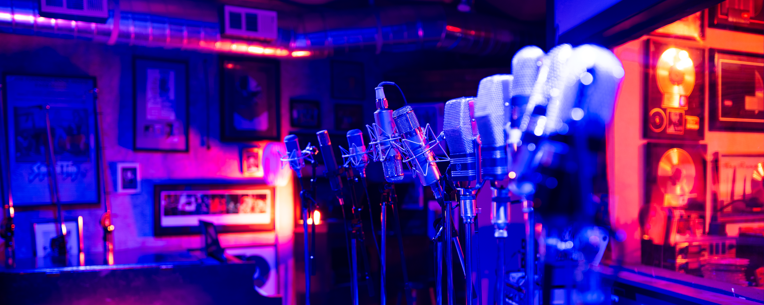 Multiple microphones on stands in a recording studio or music studio with purple and blue neon lighting, framed artwork on the walls, and audio equipment in the background.