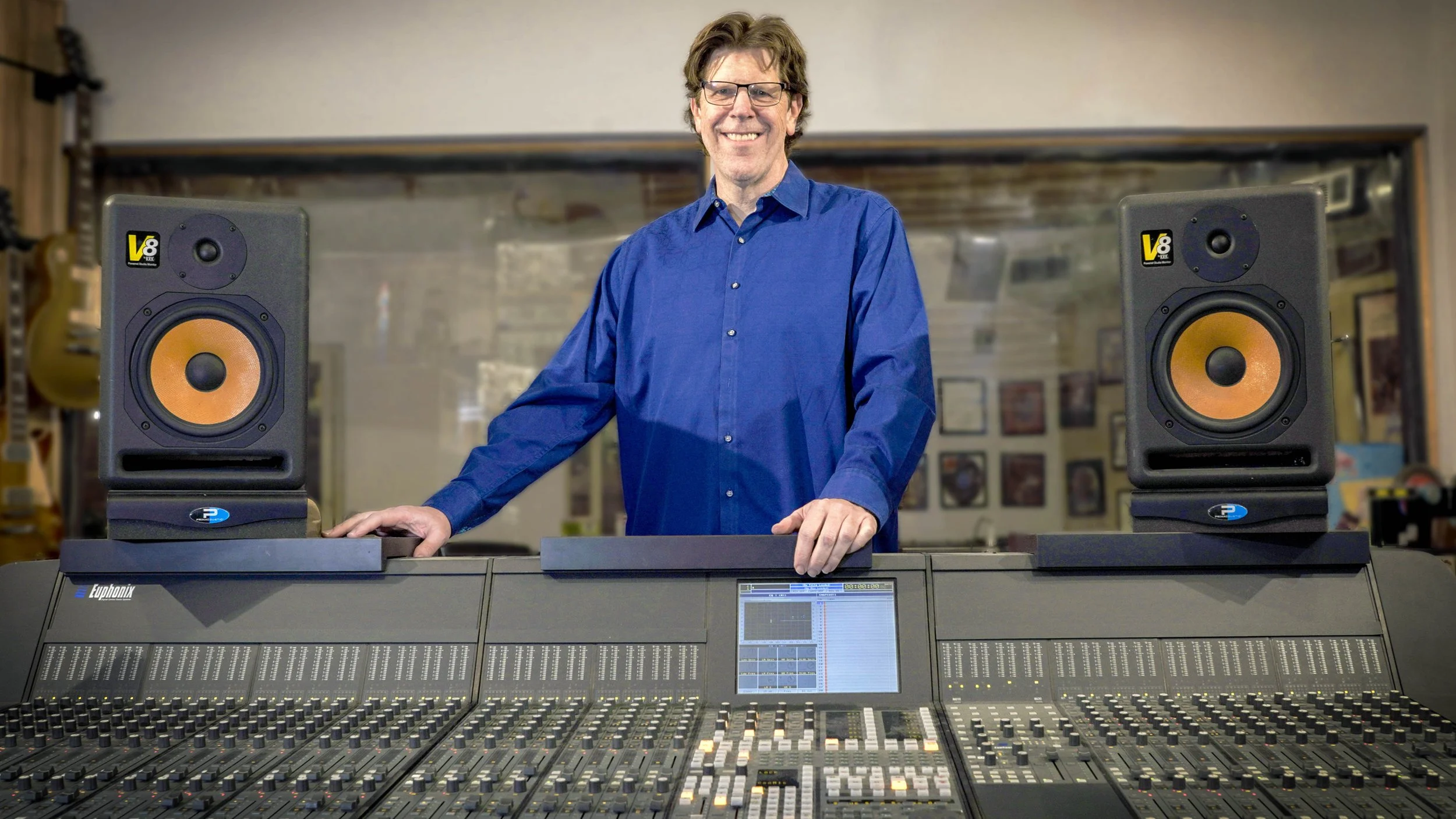 A man in a blue shirt and glasses smiling, standing behind a large audio mixing console with two studio monitors on either side.