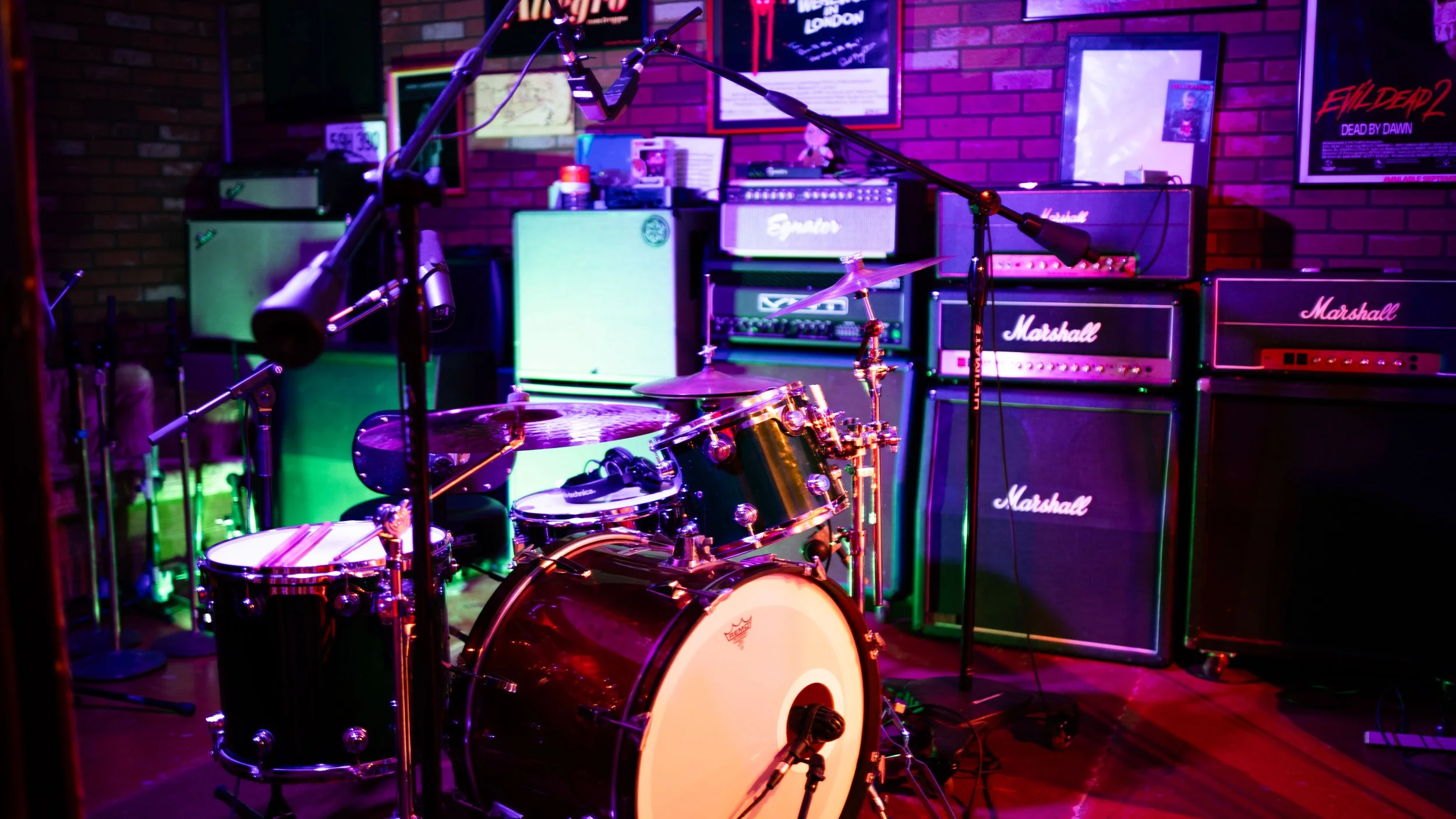 Music stage setup with a drum kit, multiple guitar amplifiers, and microphones, illuminated by colorful stage lighting in a room with brick walls.