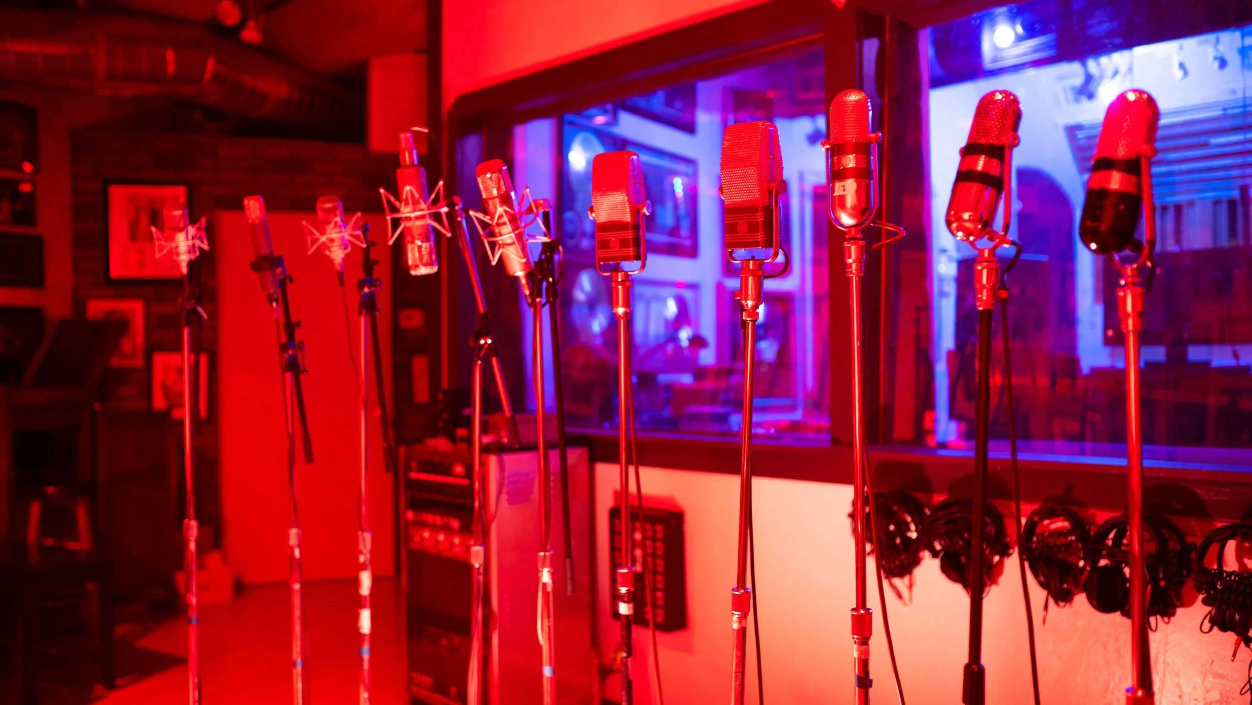 Several vintage microphones on stands inside a recording studio with red and blue lighting.