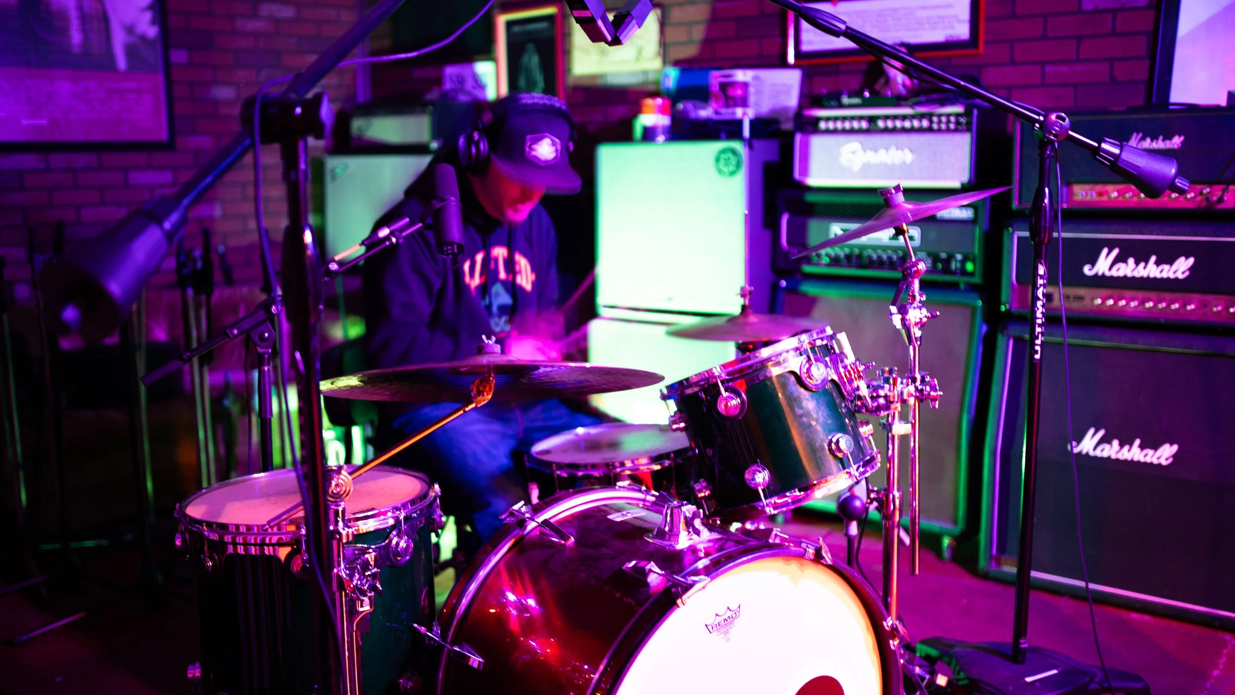 A person wearing a cap and headphones playing a drum set in a music studio with colorful lighting and multiple amplifiers in the background.