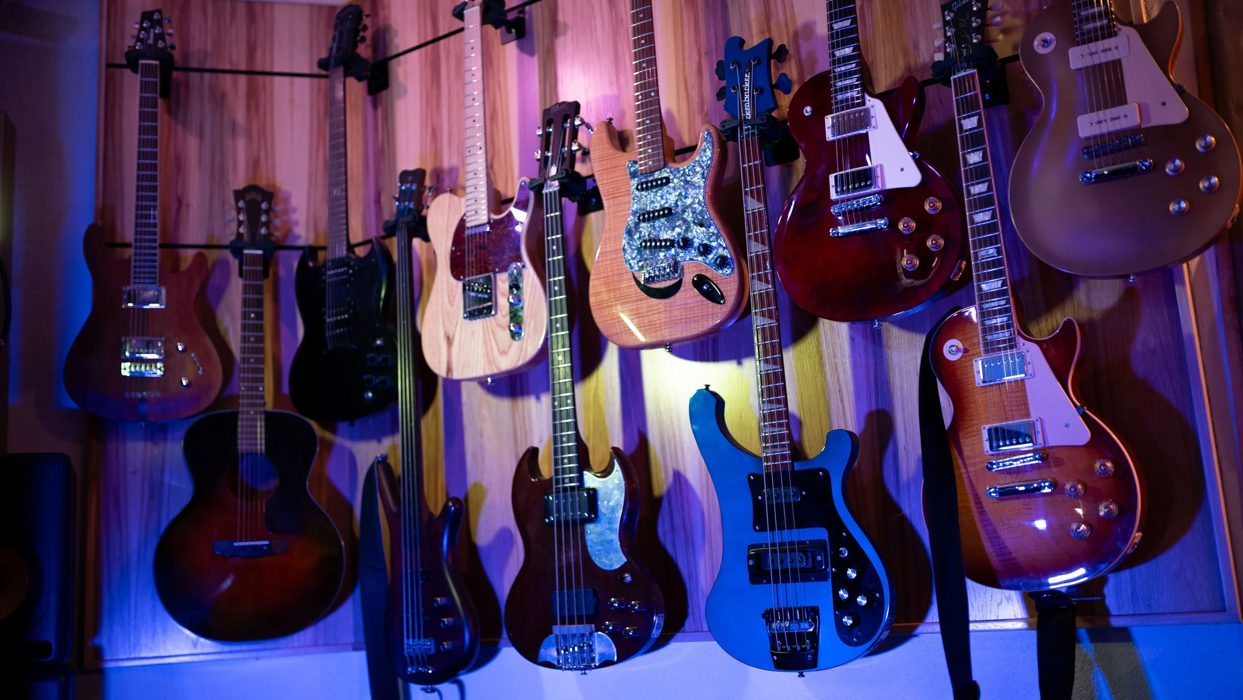 Collection of electric and acoustic guitars hanging on a wooden wall in a music studio or shop.