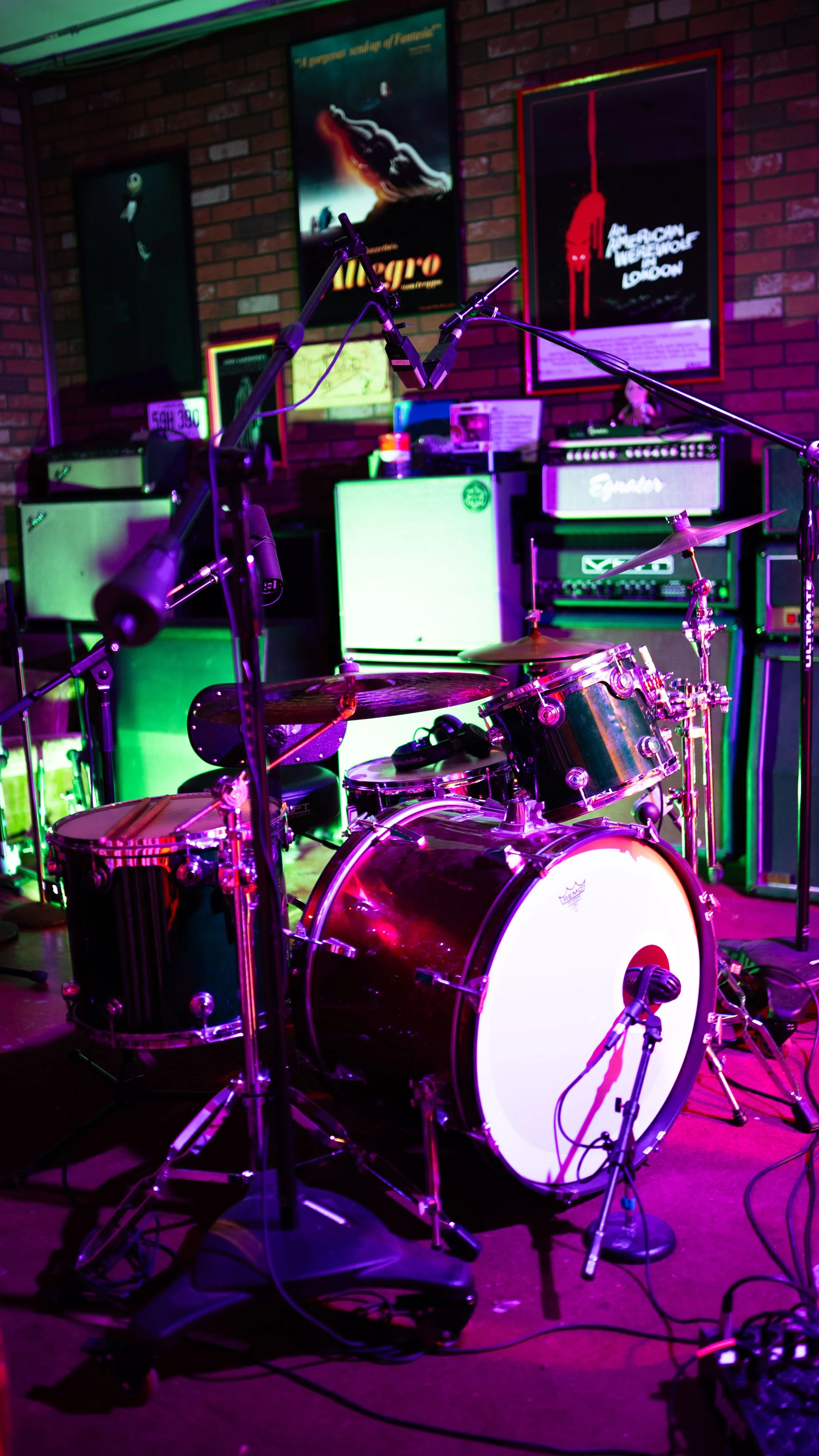 A drum set on a stage with colorful lighting, surrounded by microphone stands and musical equipment, against a brick wall decorated with vintage posters.