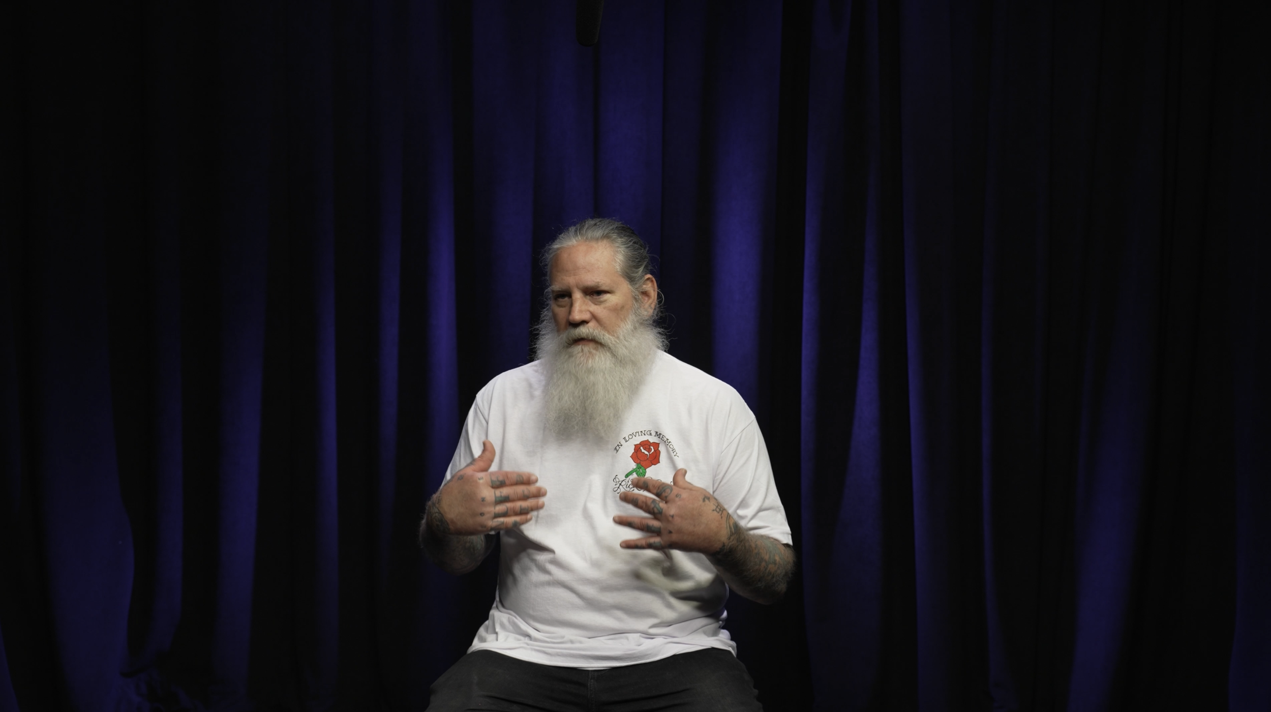 A man with long gray hair and a long white beard sitting in front of dark purple curtains, wearing a white t-shirt with a red rose graphic and text, gesturing with his hands on his chest.