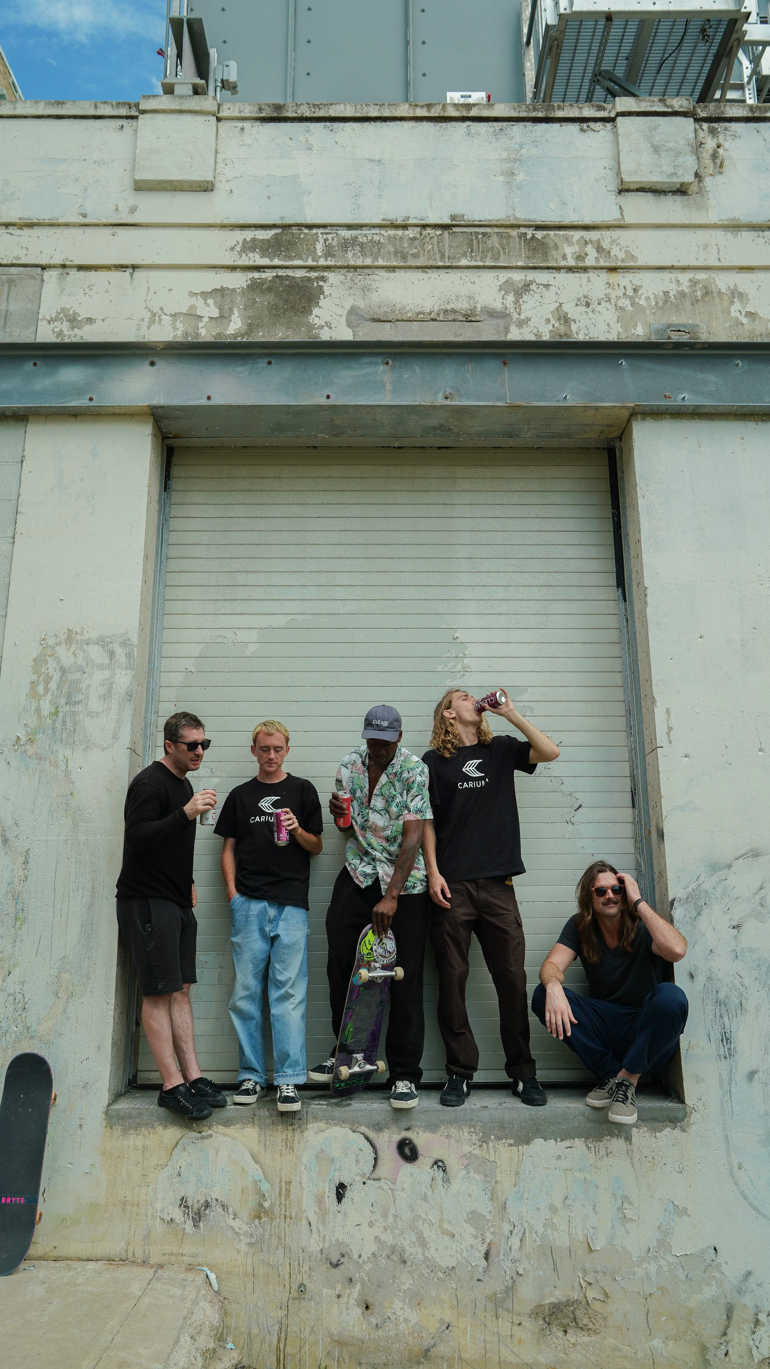 Five young men standing in front of a large, weathered garage door on a street with graffiti on the lower wall, drinking soda and holding a skateboard and a skateboard.