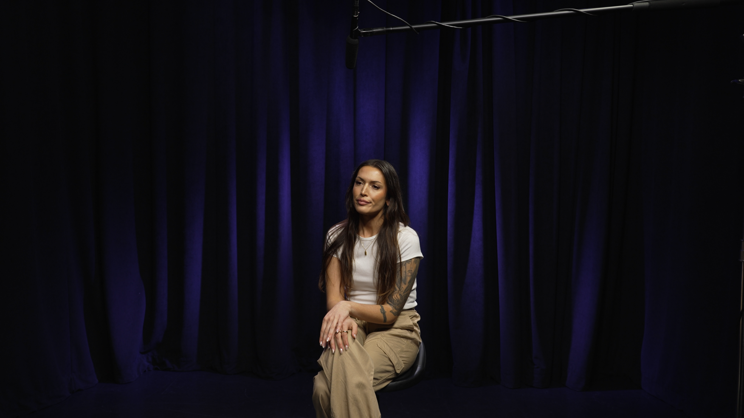 A woman with long dark hair, tattoos on her arms, sitting on a black chair in front of a blue curtain, wearing a white t-shirt and beige pants, during a interview or recording session with a microphone hanging above.