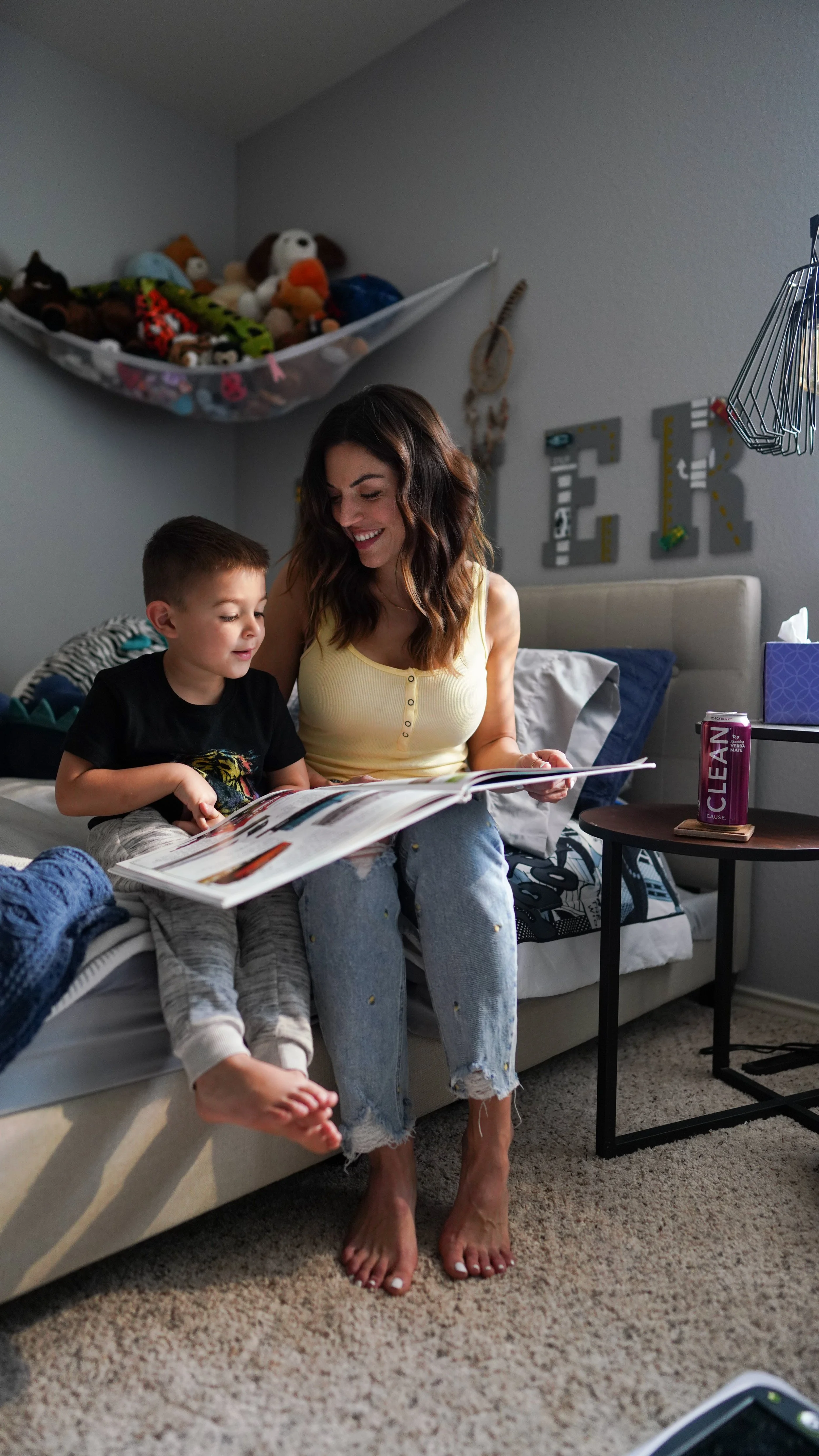 A woman and a young boy sitting on a bed, looking at a magazine together, with the boy sitting on her lap and the woman smiling. The room has a cluttered bed with clothes and a can of Lean beverage on a side table.