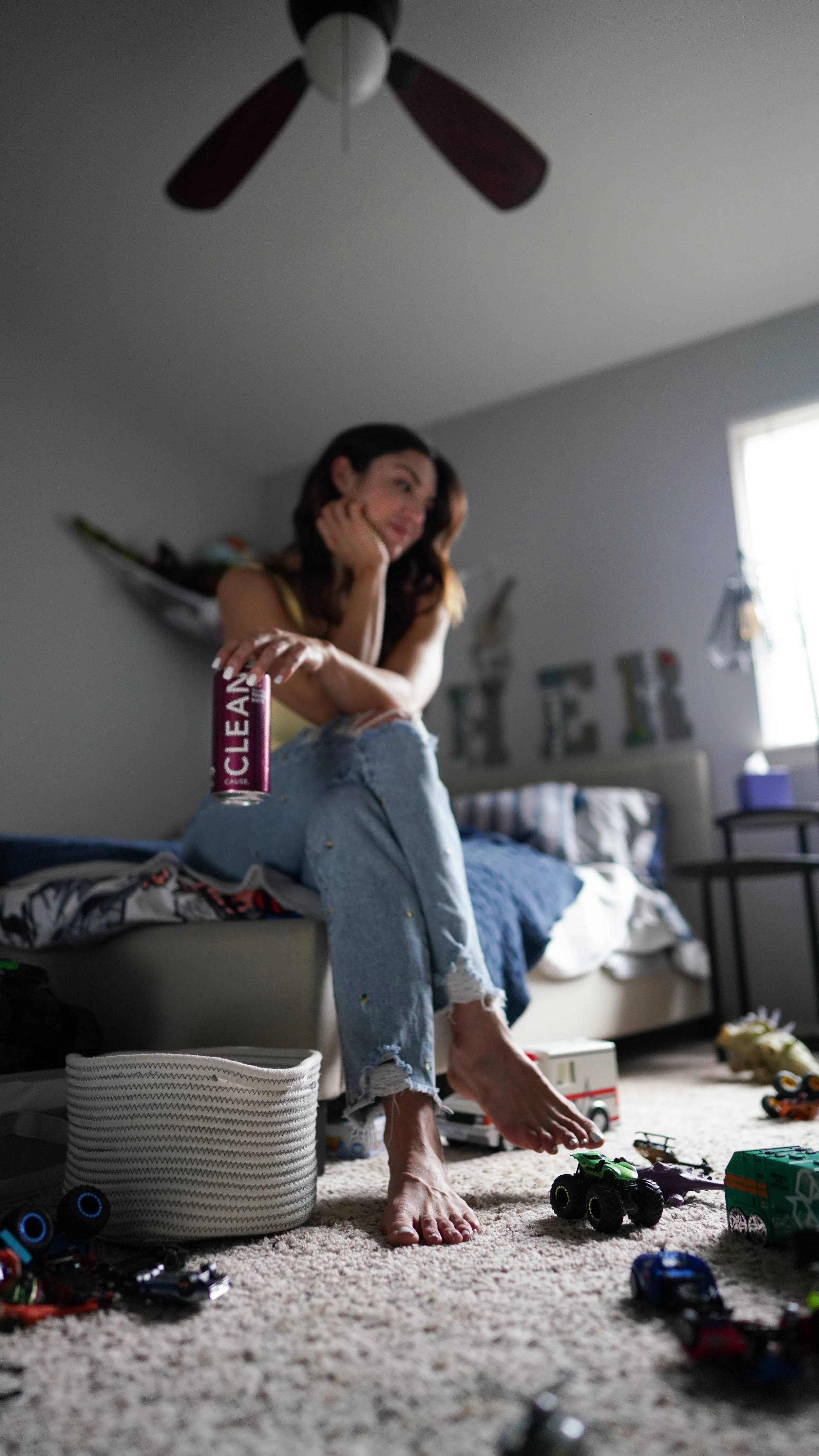 A woman sitting on a bed in a cluttered bedroom, holding a soda can, surrounded by toys and scattered items on the floor.