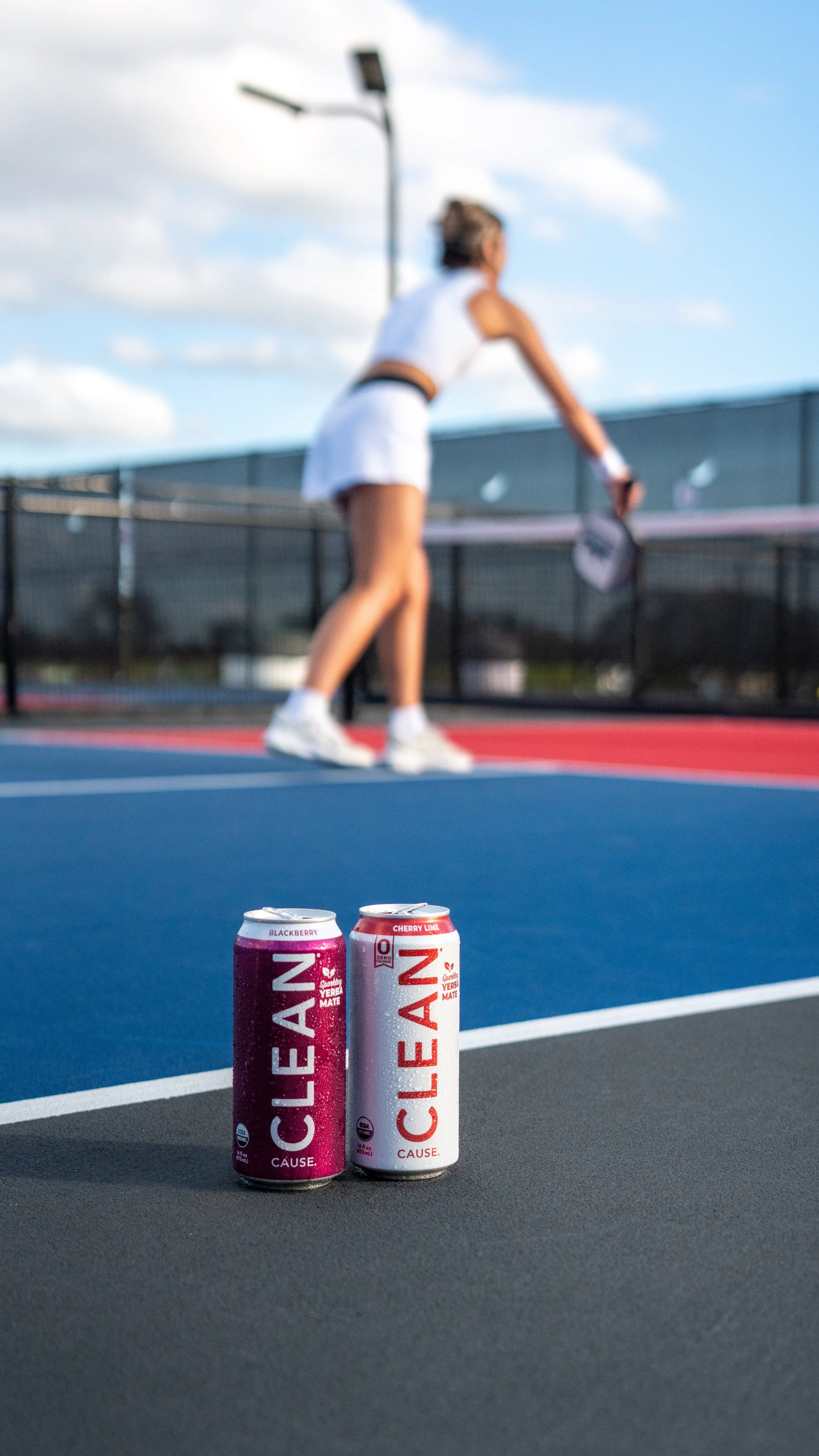 Two cans of CLEAN Cause energy drink on a tennis court with a woman playing tennis in the background.
