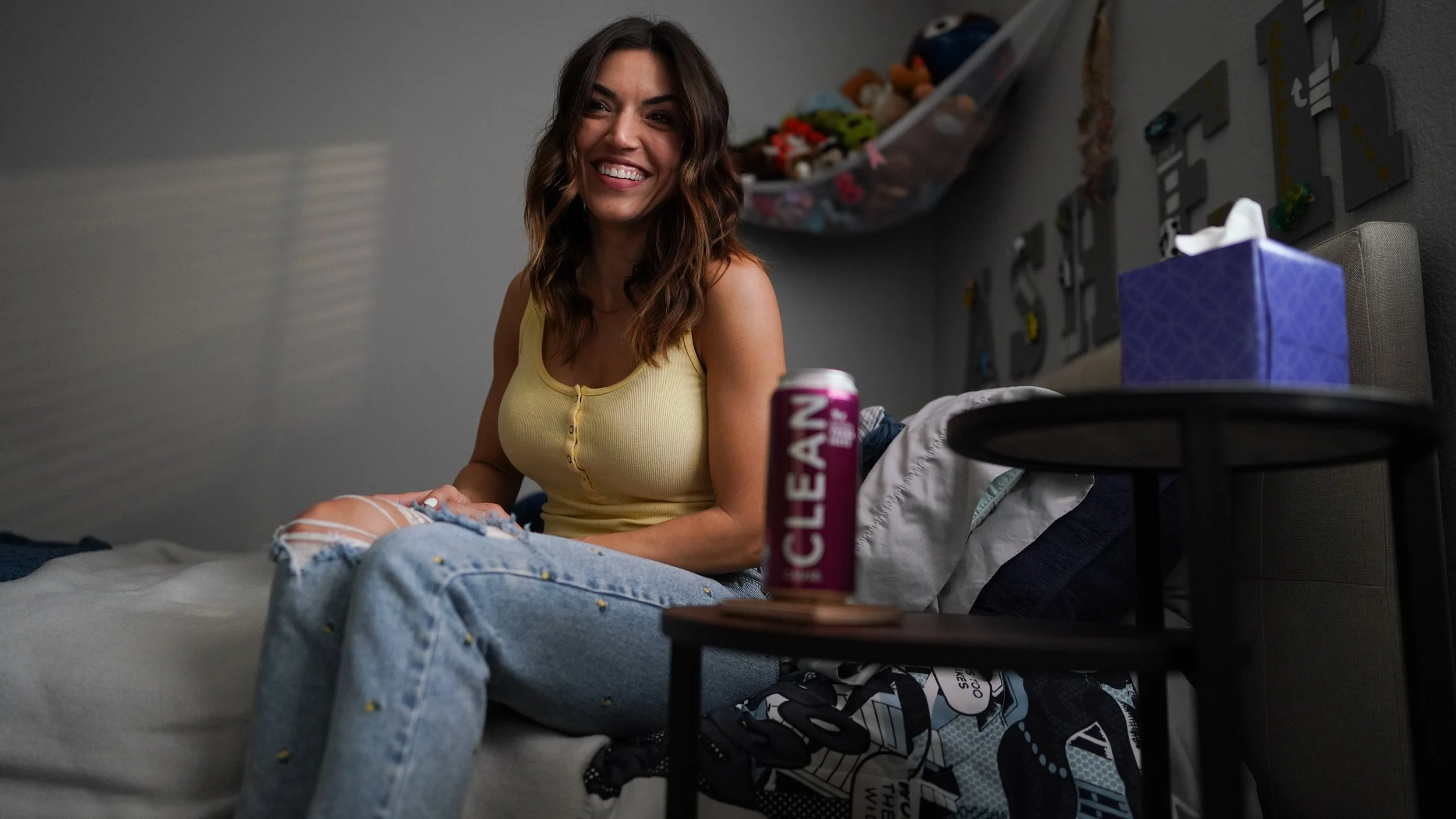 A young woman with wavy brown hair smiling and sitting on a bed in a room, with various items and toys in the background. There is a black side table with a purple box of tissue, a can of Clear brand beverage, and some clothing or fabric nearby.