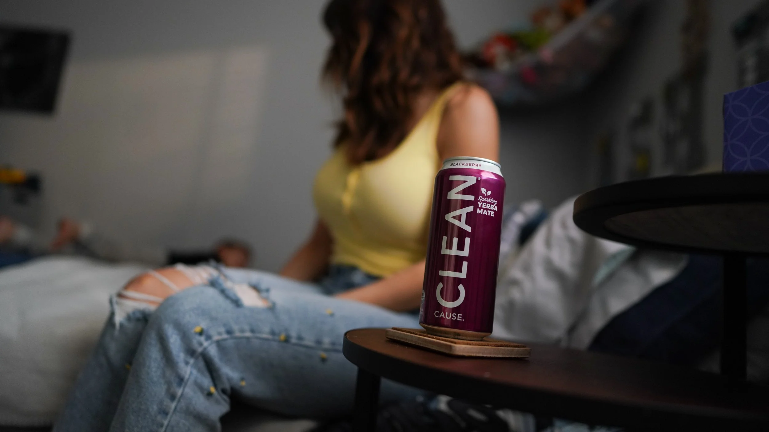 A can of ClenZeen Blackberry Yerba Mate energy drink on a table in the foreground, with a woman in a yellow top and distressed jeans sitting on a bed in the background, slightly blurred.