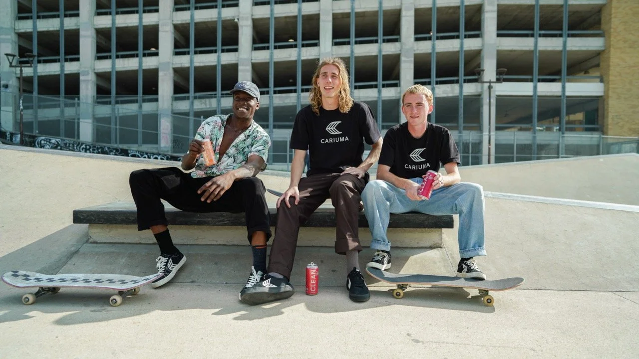 Three young men sitting on a concrete ledge at a skate park, holding drinks and skateboards, with a building in the background.