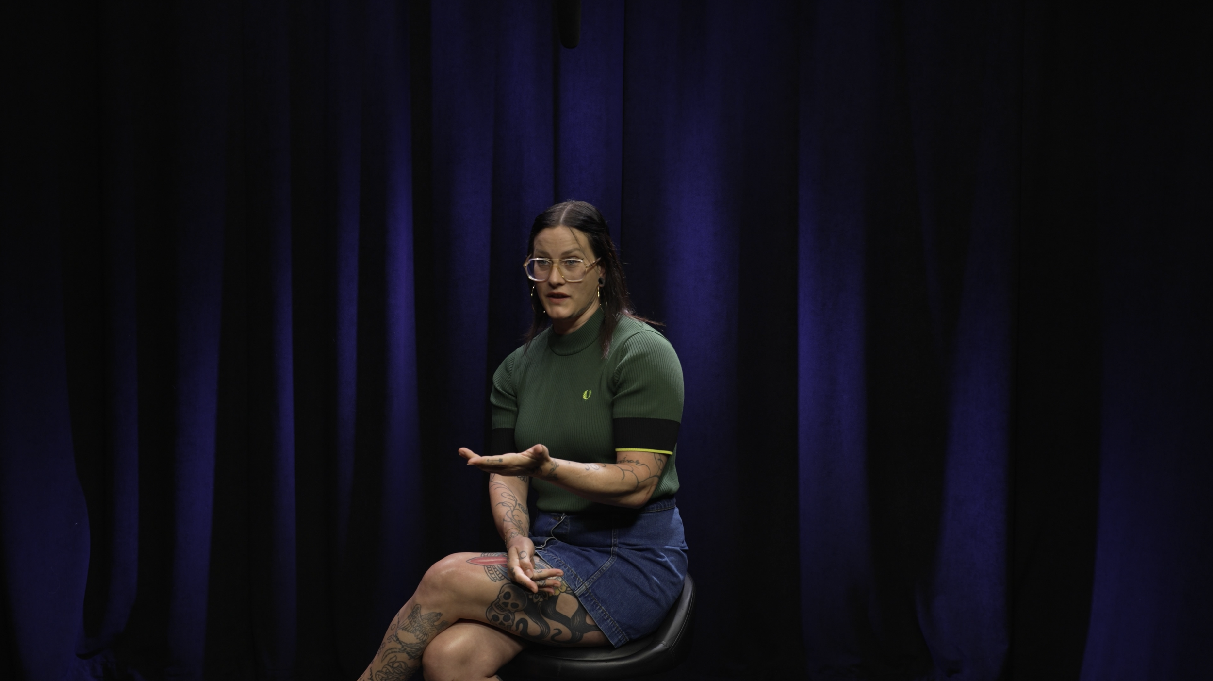A woman with glasses and tattoos, wearing a green shirt and denim skirt, sitting on a chair against a dark blue curtain background.