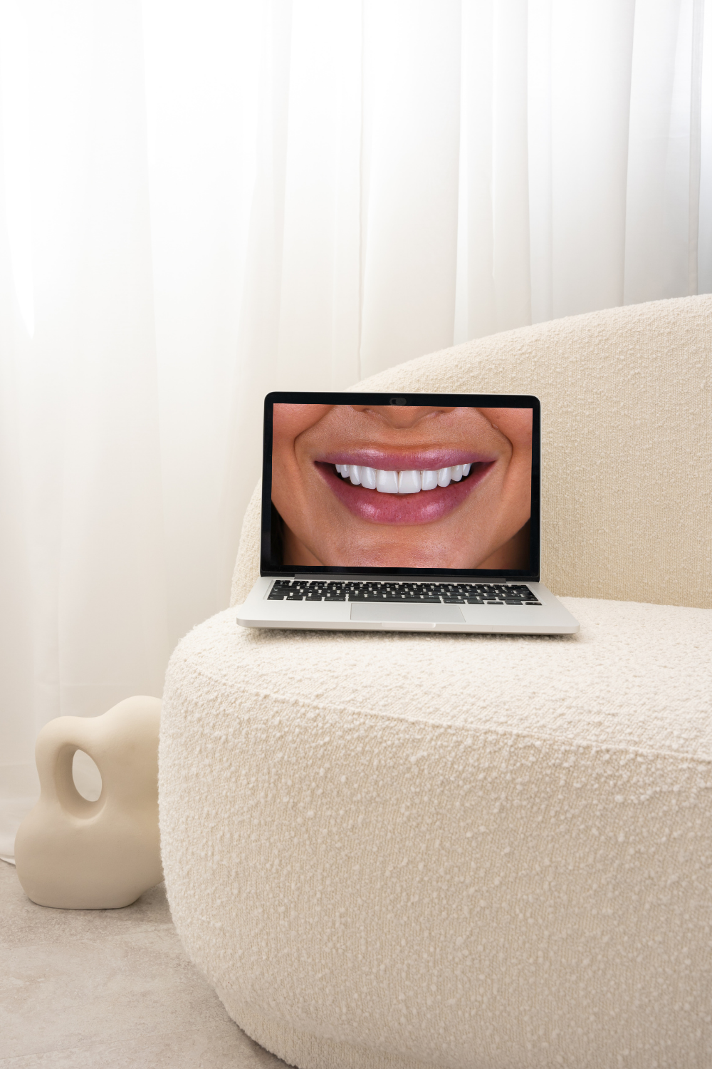 Laptop on beige textured armchair displaying a close-up of a smiling person's mouth with white teeth.