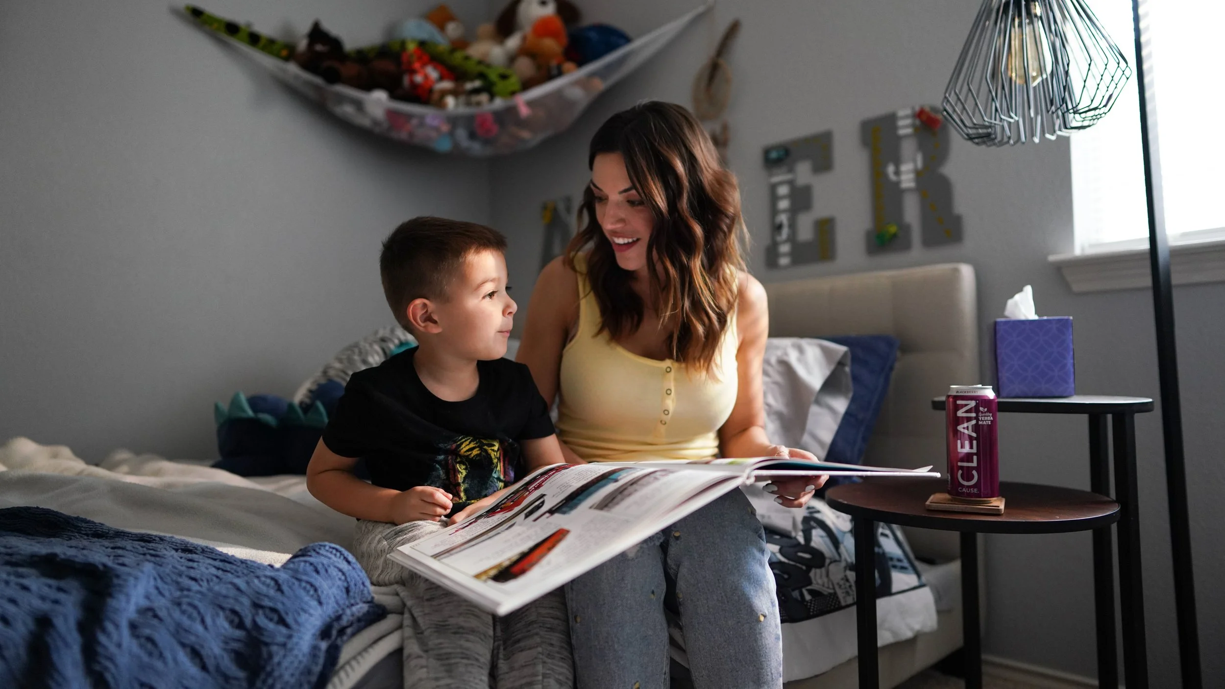 A woman and young boy sitting on a bed reading a magazine together in a bedroom. There are plush toys on a shelf above them, a tissue box, and a soda can on the side table.