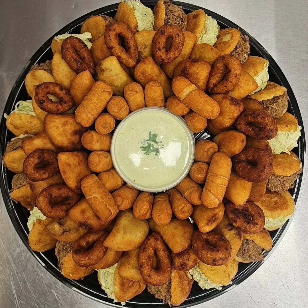 A large round platter of assorted fried foods surrounding a cup of green dipping sauce, including onion rings, fried chicken pieces, spring rolls, fried plantains, fried potato wedges, and other fried snacks.