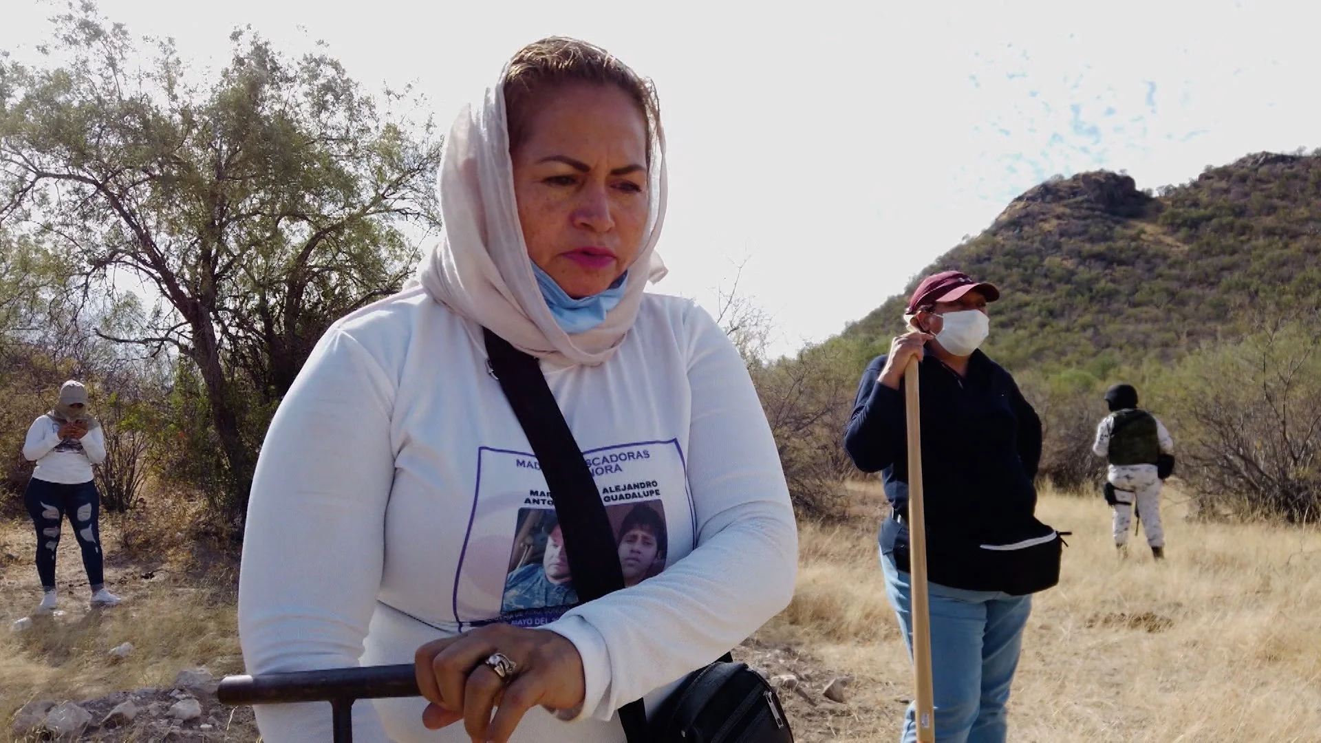Grupo de personas en un área rural con vegetación seca. Una mujer al frente usa un pañuelo en la cabeza y lleva una camiseta con una foto impresa. Otra persona sostiene un palo y está cubierta con una mascarilla, mientras unos arbustos y montañas se ven al fondo.
