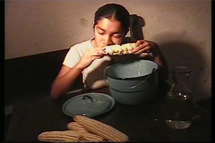 Persona comiendo maíz hervido de una cubeta, con una jarra de agua en la mesa.
