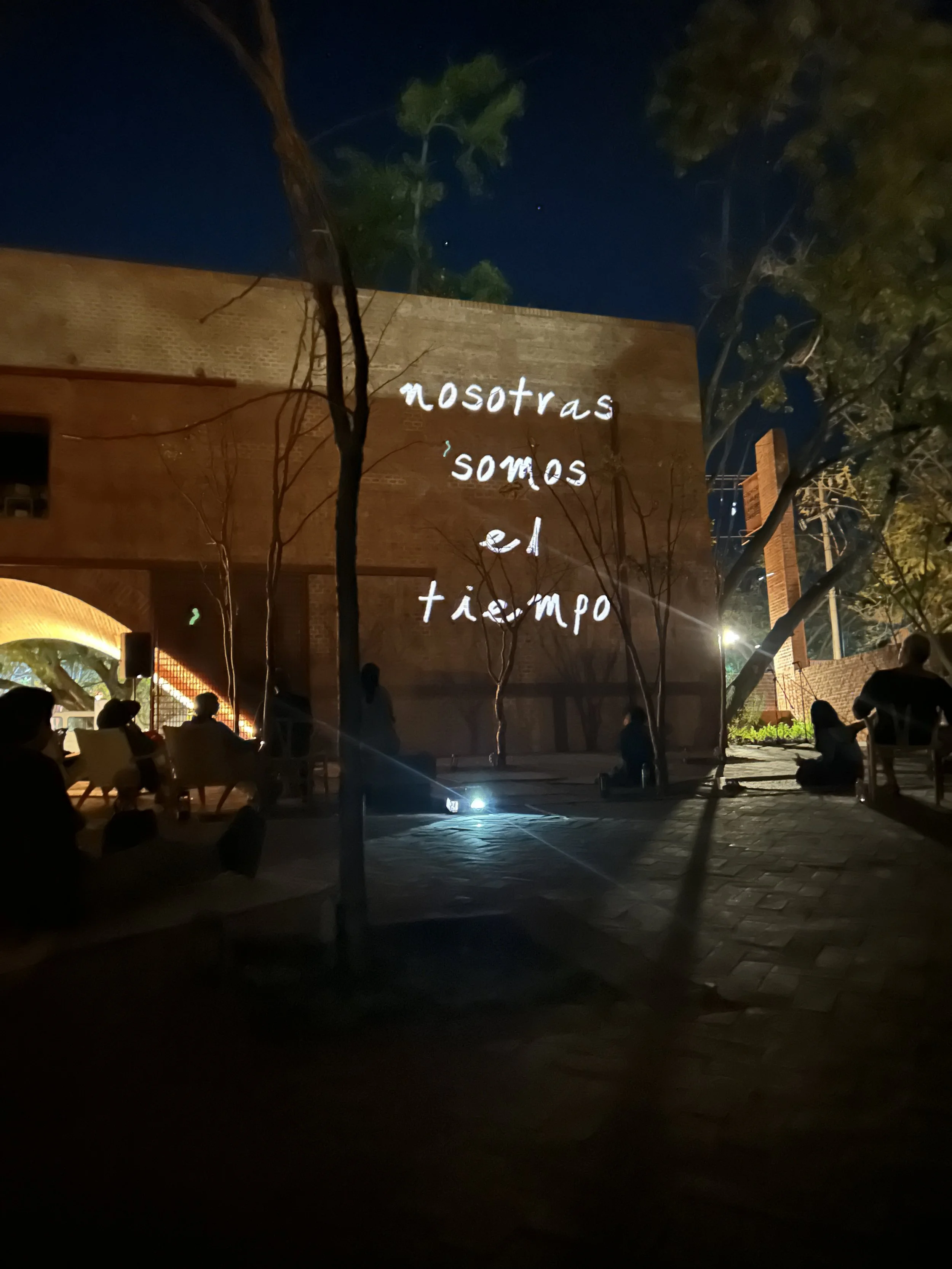 Proyección de las palabras "nosotras somos el tiempo" en la pared de un edificio de ladrillo. Se observa un patio con árboles y siluetas de personas sentadas durante la noche.