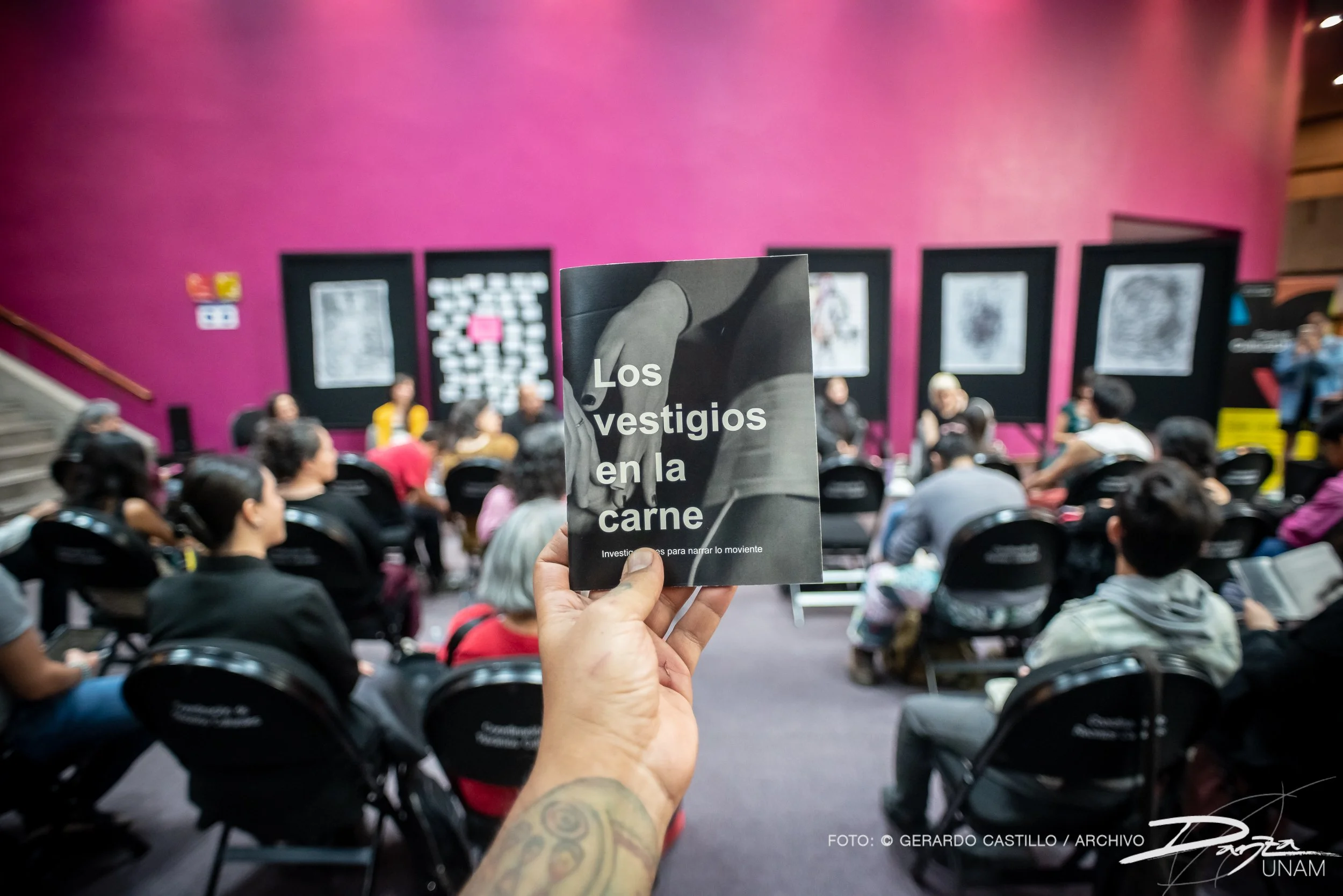 Persona sosteniendo un folleto titulado "Los vestigios en la carne" en un evento cultural con personas sentadas en un entorno interior con una pared rosa de fondo.
