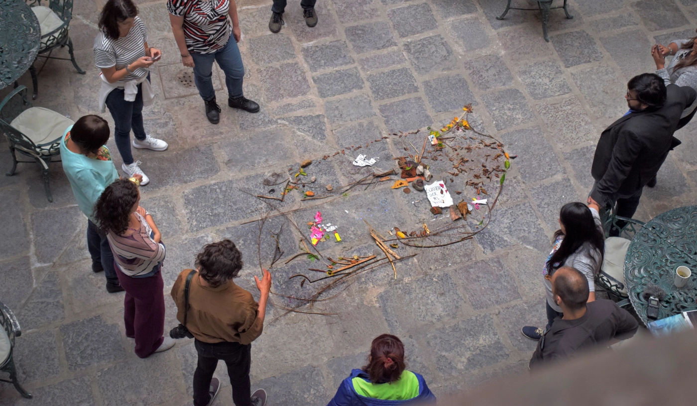 Grupo de personas formando un círculo alrededor de un altar improvisado en el suelo, decorado con ramas, hojas, y papeles, en un patio de piedra.