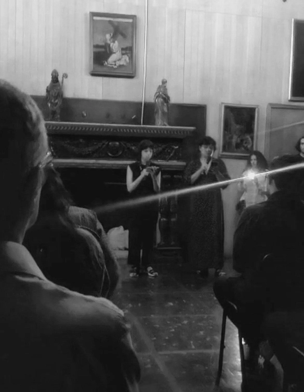 Varias personas en una sala antigua con cuadros y estatuas; una mujer está hablando frente a la audiencia.