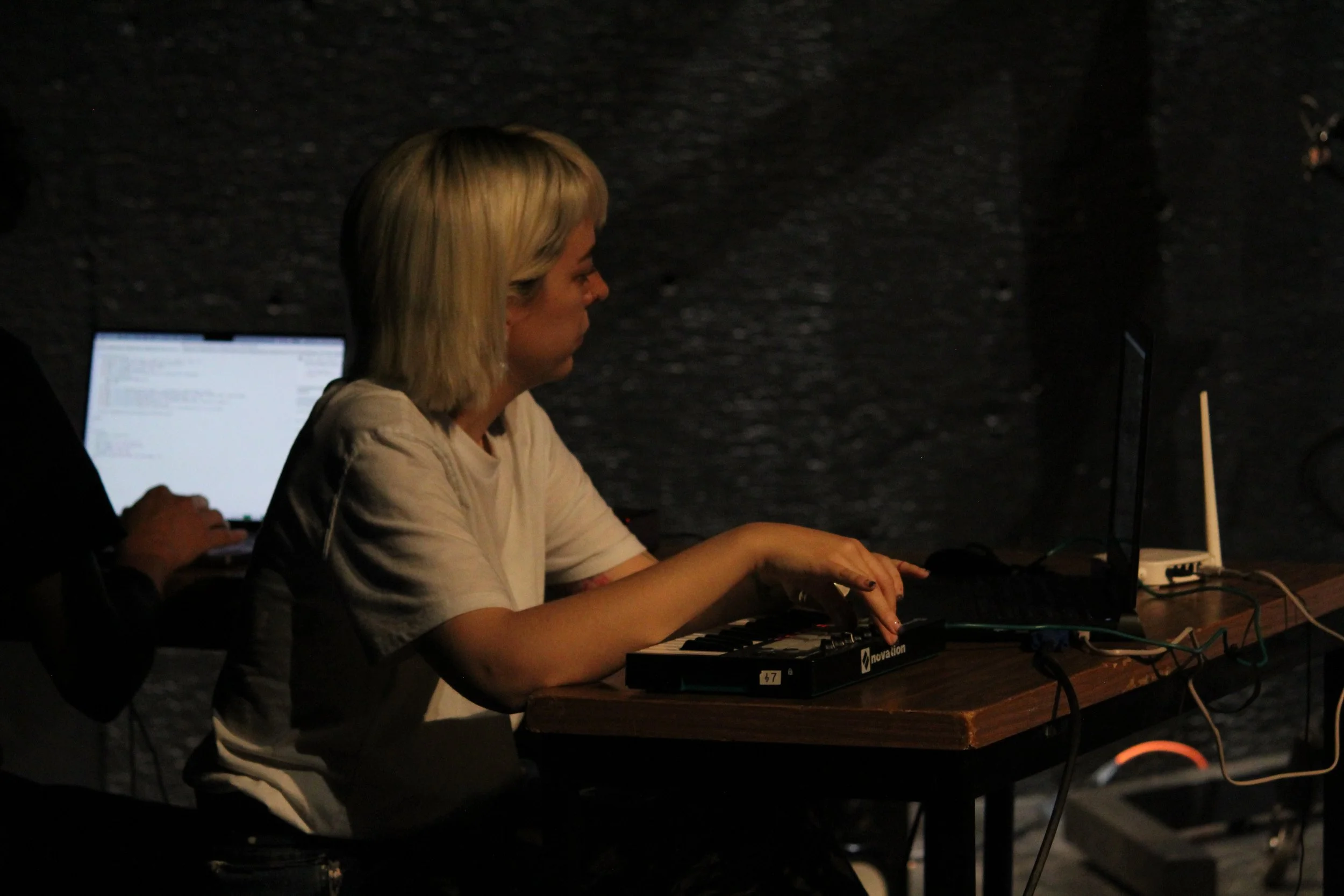 Dos personas trabajando en una mesa con computadoras portátiles y un teclado MIDI, en un entorno oscuro.