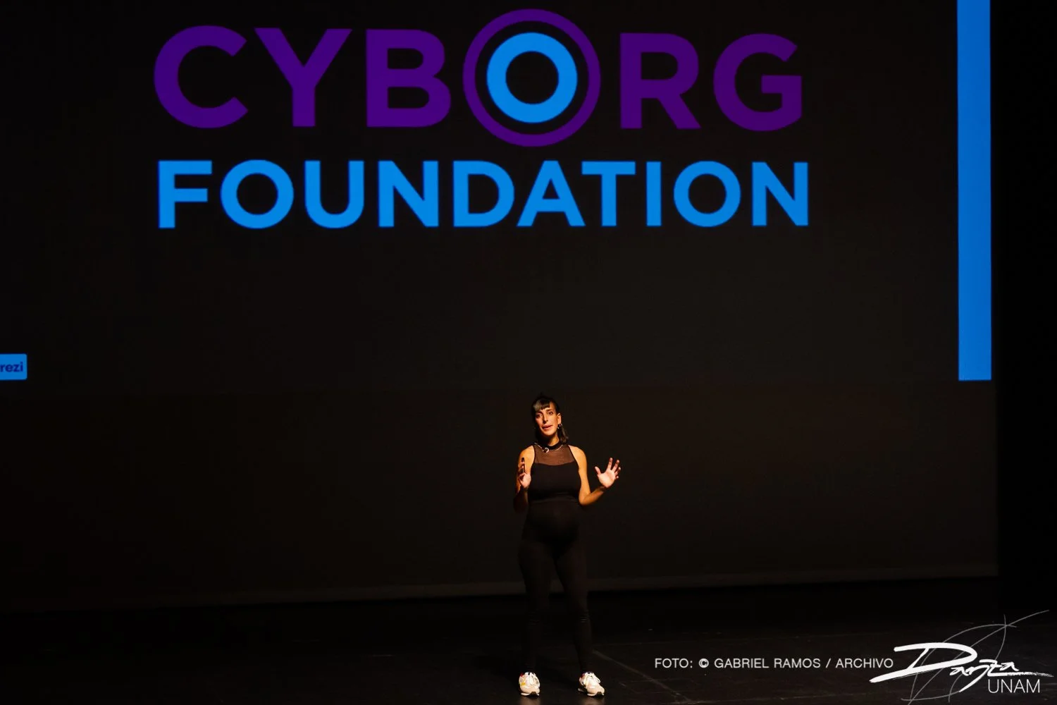 Una persona en el escenario hablando frente a un proyector que muestra el texto "Cyborg Foundation" en letras grandes. El fondo es oscuro y la persona lleva ropa negra.