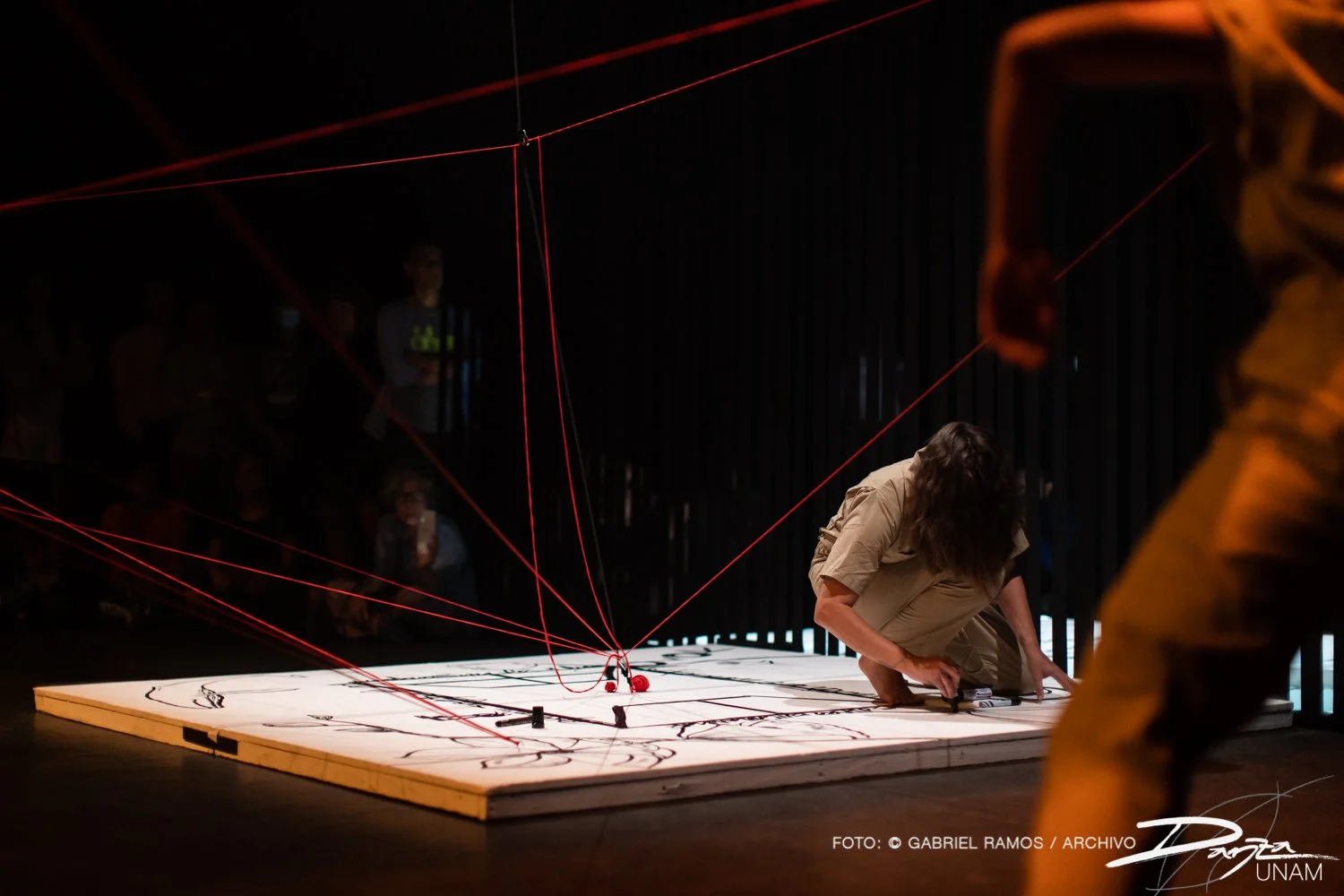 Instalación artística con cuerdas rojas y una persona en el suelo dibujando en un panel blanco, espectadores al fondo.