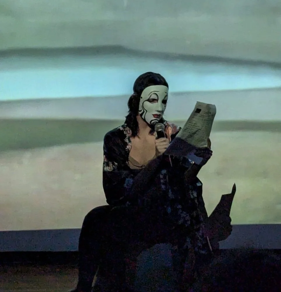 Persona con máscara artística leyendo un papel en un escenario con fondo proyectado.