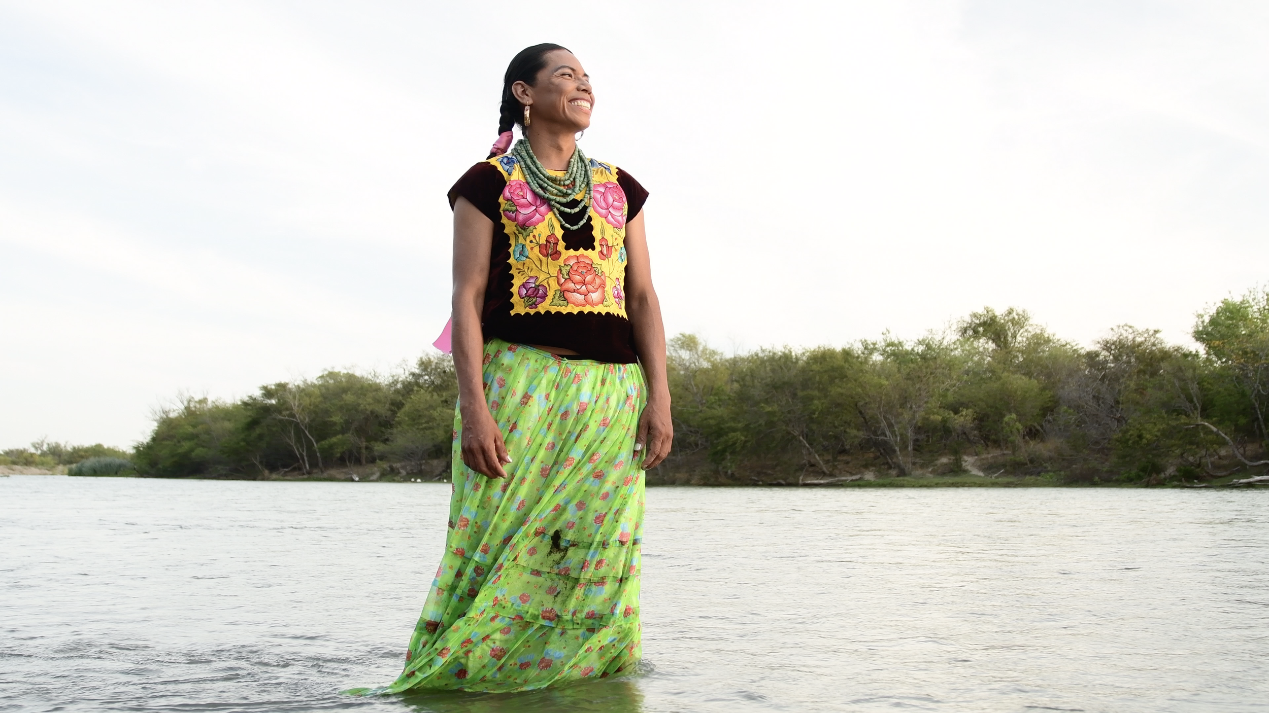 Mujer con vestido tradicional mexicano de pie en un río, fondo de vegetación verde.
