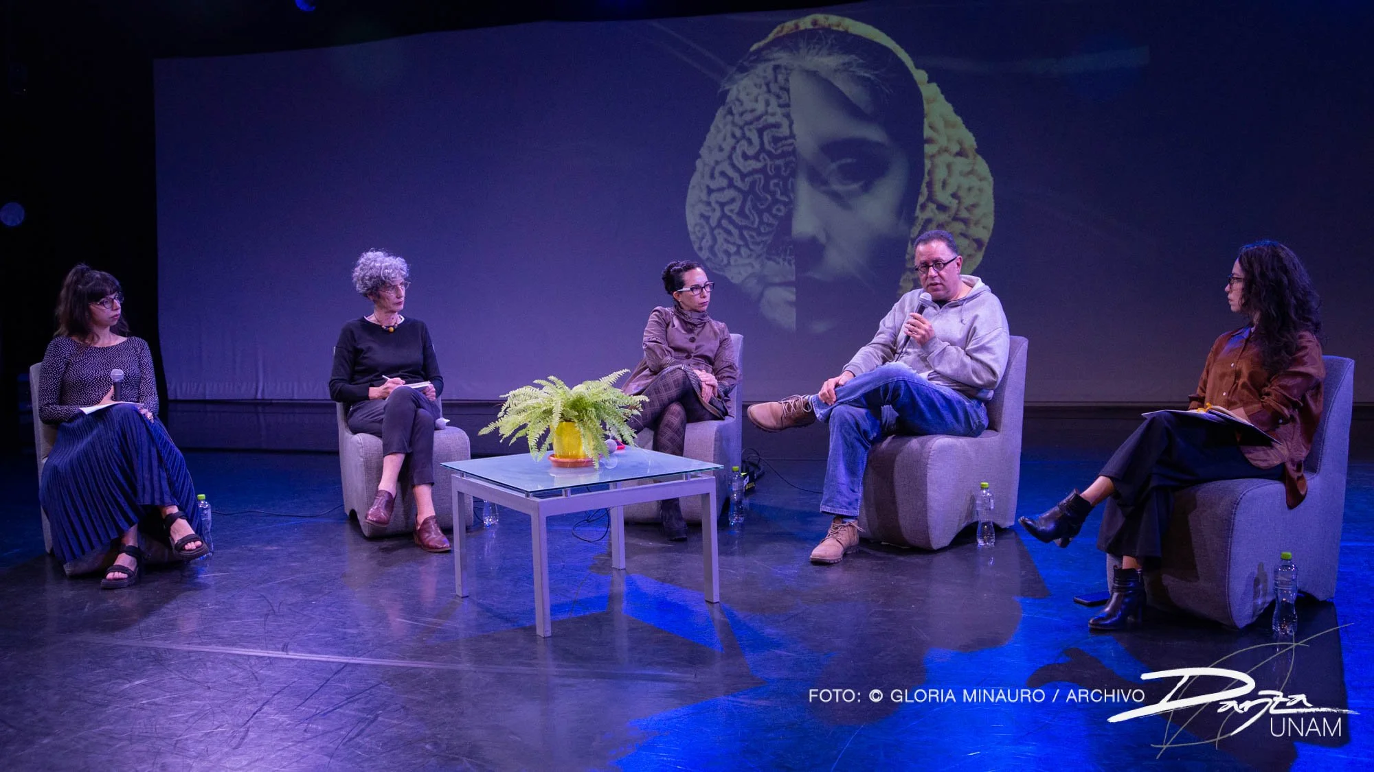 Cinco personas sentadas en un escenario participando en un panel de discusión. Hay una mesa con una planta en el centro y botellas de agua en el suelo. De fondo, una imagen de una cara humana y un cerebro proyectados en una pantalla.