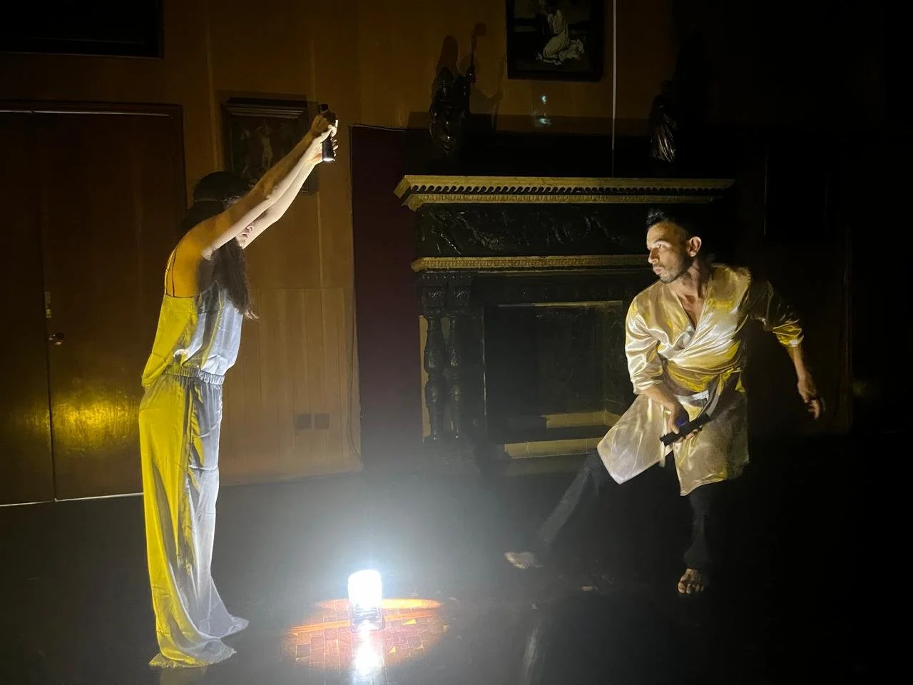 Dos personas en una performance dramatizada en una habitación con luz tenue; una sostiene una botella hacia arriba y el otro adopta una postura dinámica.