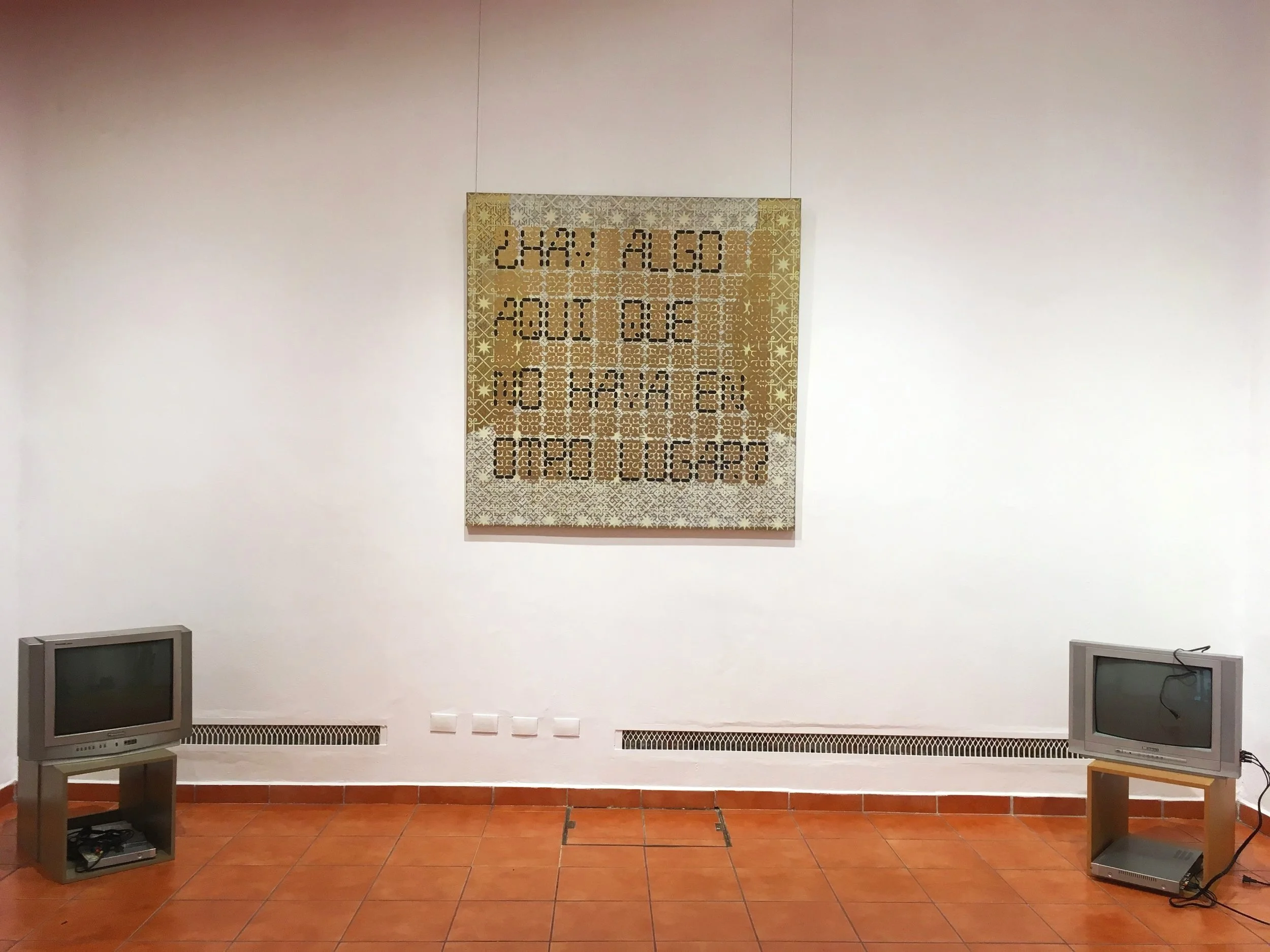 Dos televisores antiguos en un museo, con una pieza de arte colgada en la pared entre ellos. El arte tiene patrones geométricos y texto que parece español.