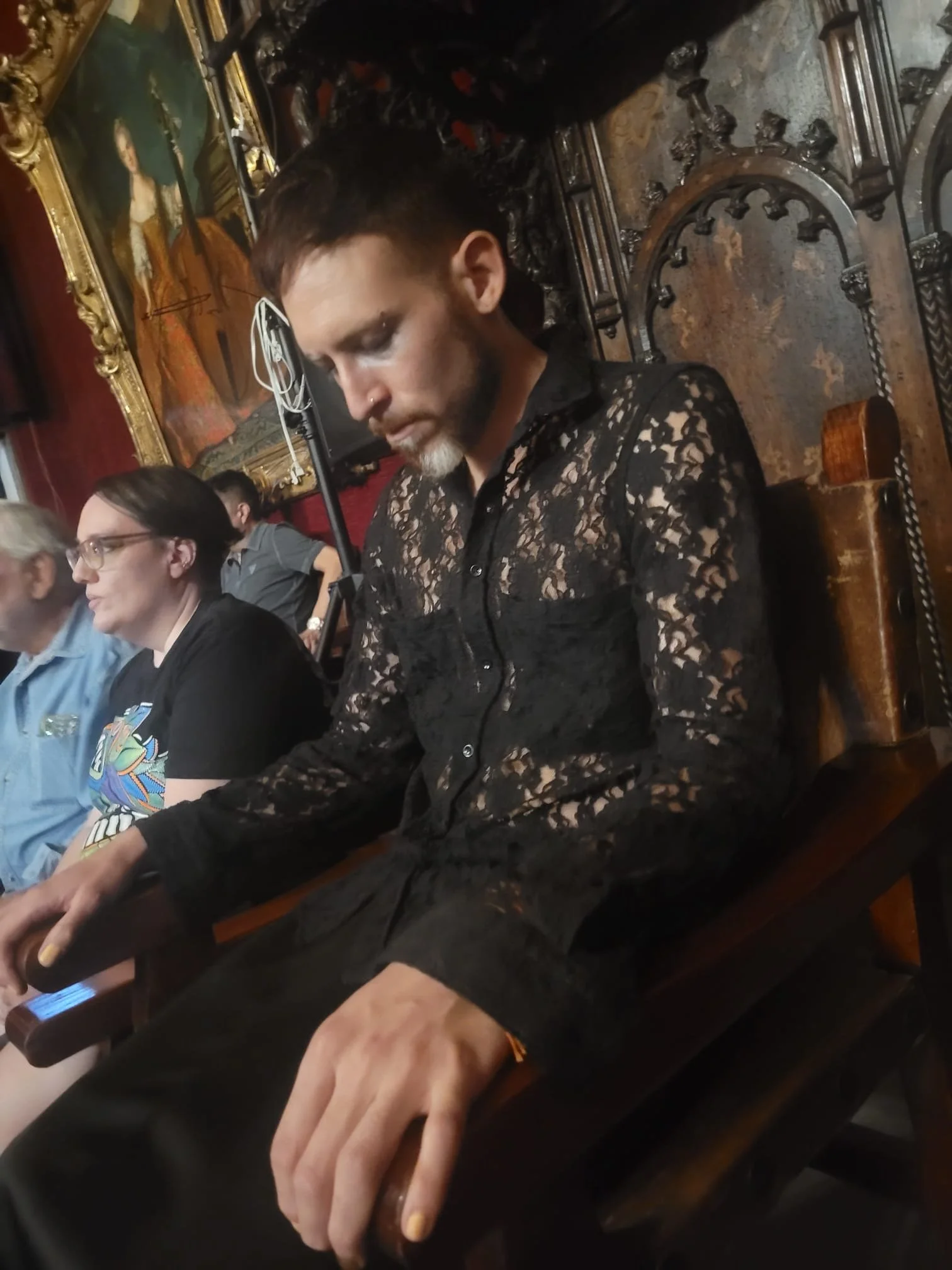 Persona con camisa de encaje negro, sentada en silla de madera tallada, rodeada de otras personas, con un cuadro al fondo.