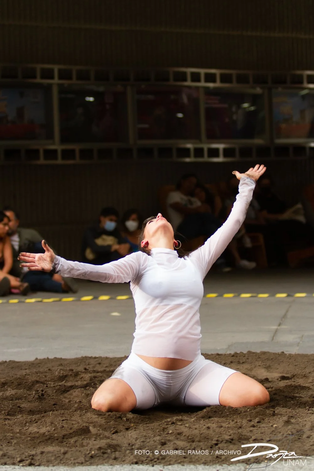 Persona en performance, arrodillada sobre tierra, con brazos extendidos y mirada hacia arriba, vestida de blanco, en un espacio público con público de fondo.